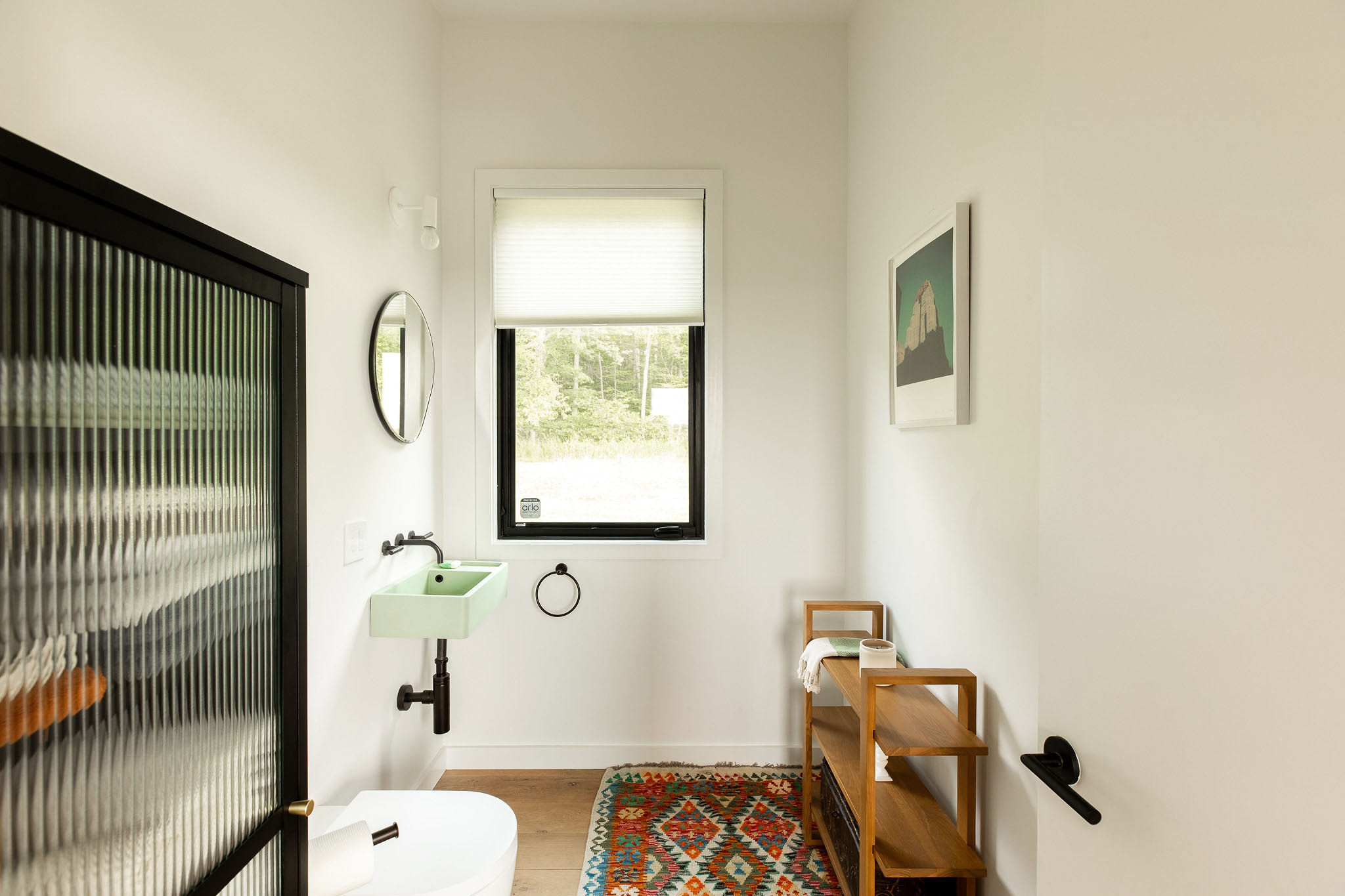 Compact powder room with a unique light green wall-mounted sink and a black-framed fluted glass door, featuring a small window and a colorful patterned rug on hardwood floors.