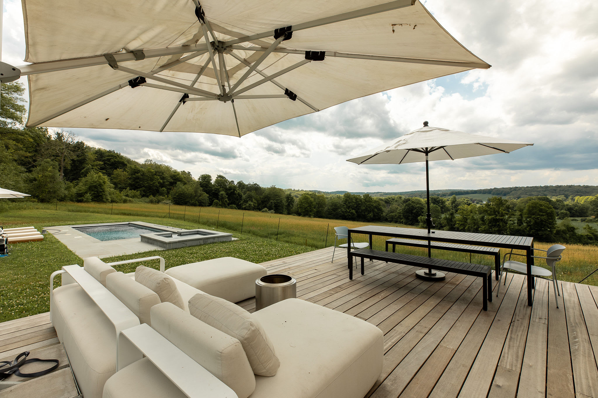 Expansive wooden deck furnished with a large white outdoor sectional sofa and a dining table with benches under a wide white umbrella. The swimming pool and hot tub are visible in the grassy area beyond the deck, surrounded by lush green landscape.