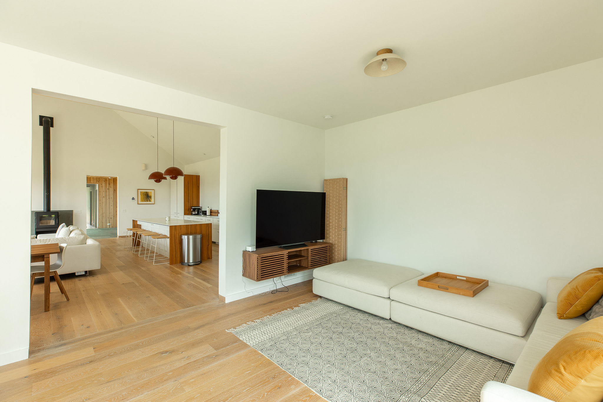 Bright living room with a large sectional sofa, a wall-mounted TV on a wooden console, and a patterned rug, offering an open view into a modern kitchen and dining area with a wood-burning stove.