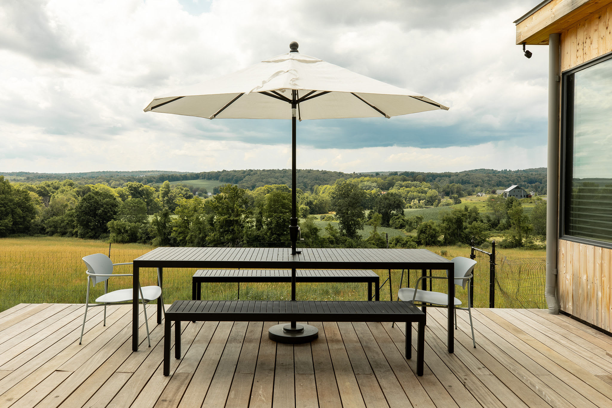 Outdoor dining area on a wooden deck, featuring a long black dining table with matching benches and chairs, shaded by a large white umbrella. The deck overlooks a vast green landscape with rolling hills and distant trees under a cloudy sky.