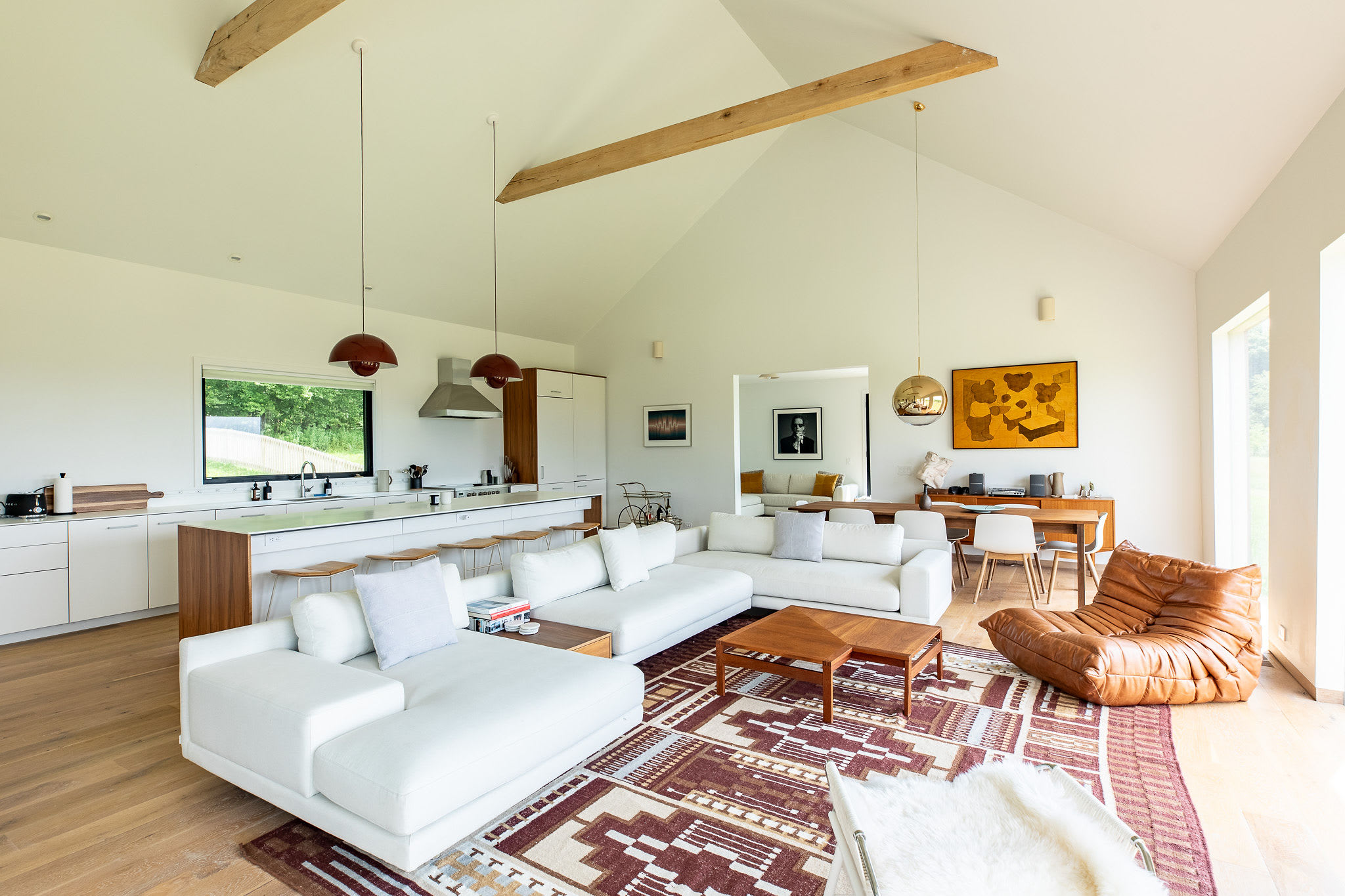 An open-concept living area features two large white sectional sofas on a patterned rug, facing a modern kitchen with a long island. The room boasts vaulted ceilings with exposed wooden beams and a mix of contemporary and mid-century modern furniture.