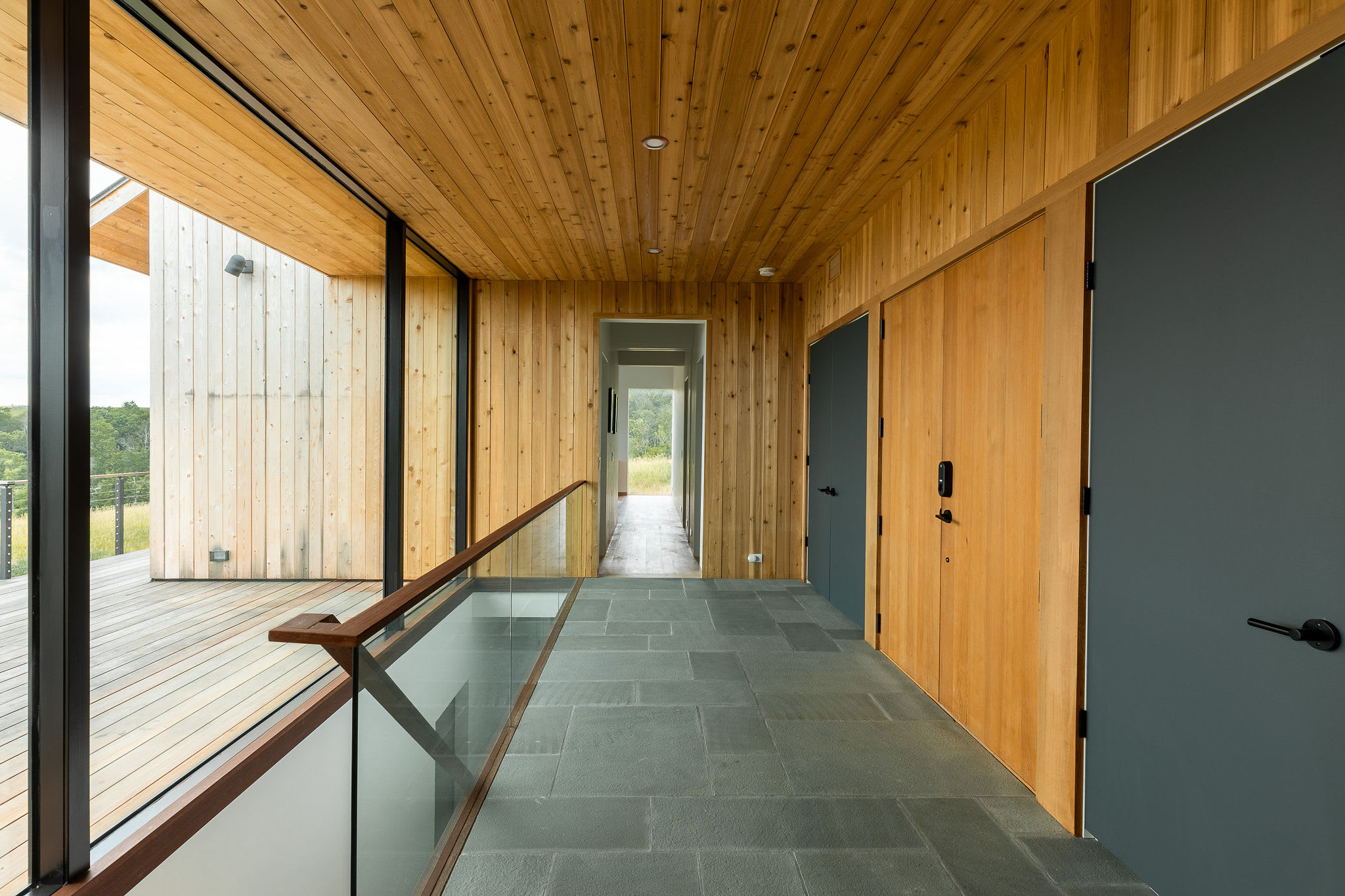 A modern interior hallway features warm wooden paneling on walls and ceiling, contrasting with dark grey slate tile flooring. A glass railing overlooks an outdoor deck, while dark doors line one side of the hall.