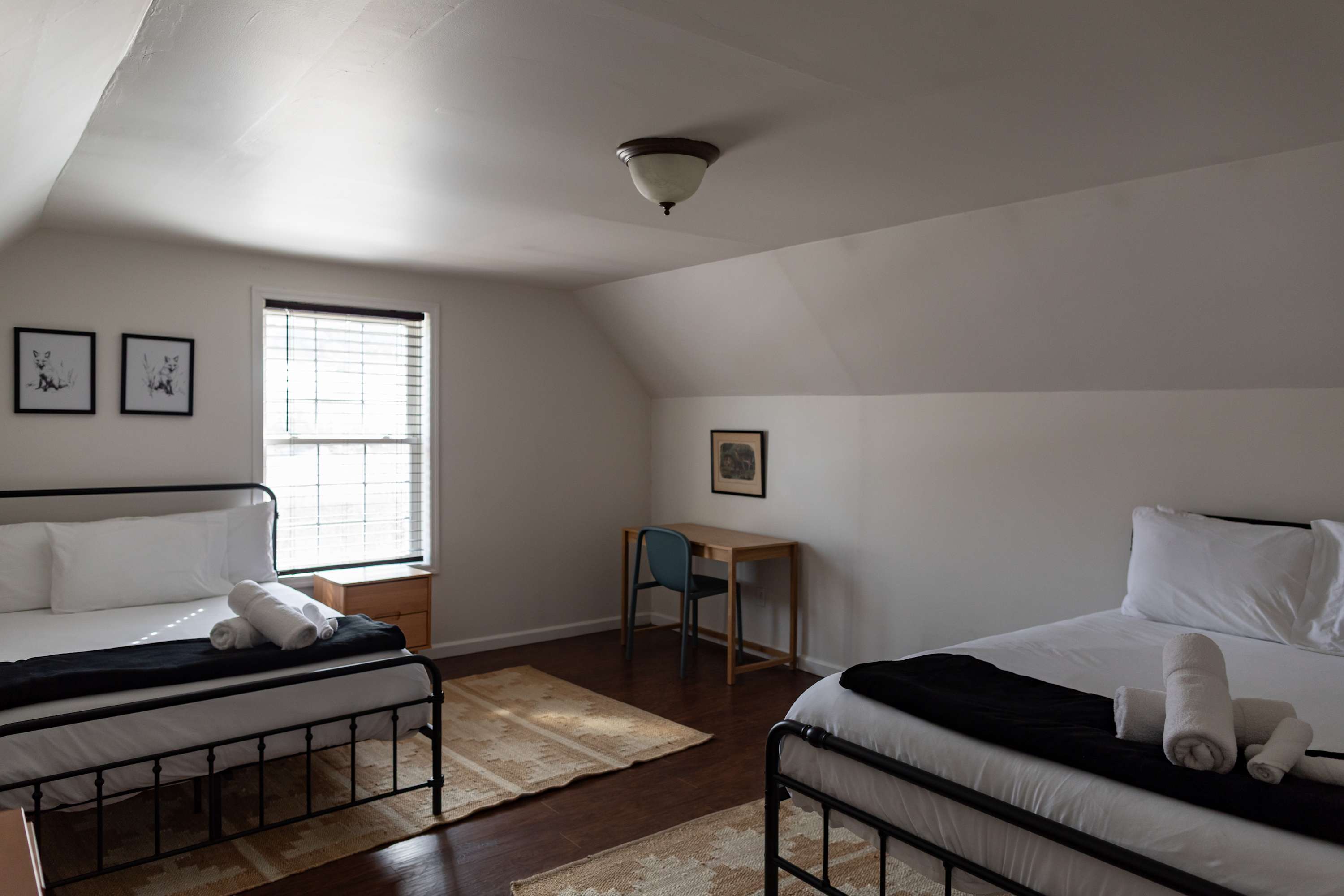 A spacious bedroom featuring two black metal beds with white bedding and black throws, set under a sloped ceiling. Two framed fox prints adorn the wall, and a wooden desk with a chair is visible in the corner.