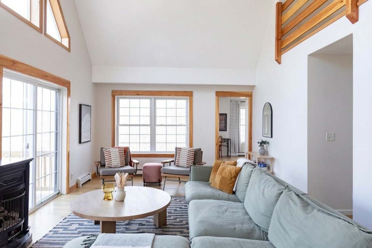 A bright and airy living room featuring a large light green sectional sofa, two gray armchairs, a round wooden coffee table, and light wood trim around windows and sliding glass doors, with a patterned rug grounding the space.