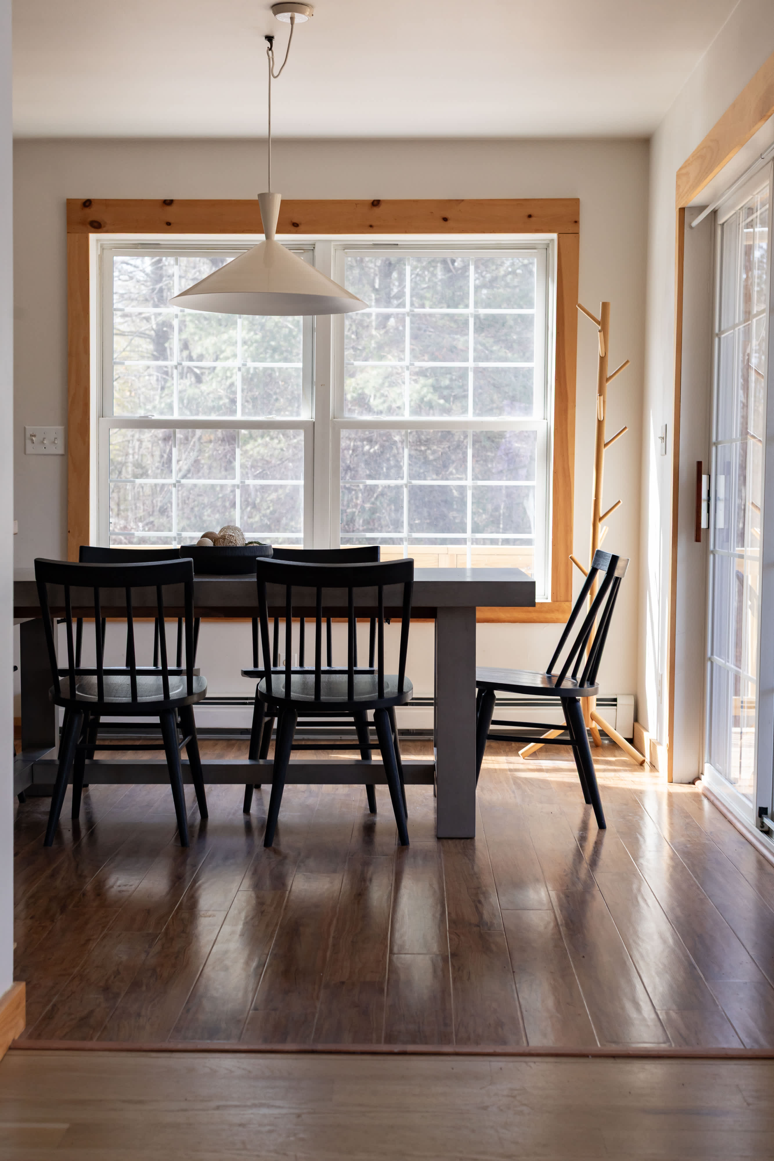 A bright dining area features a large grey rectangular table with four black spindle-back chairs, illuminated by a modern white pendant light. Large windows with white grids and natural wood trim overlook an outdoor deck and trees.