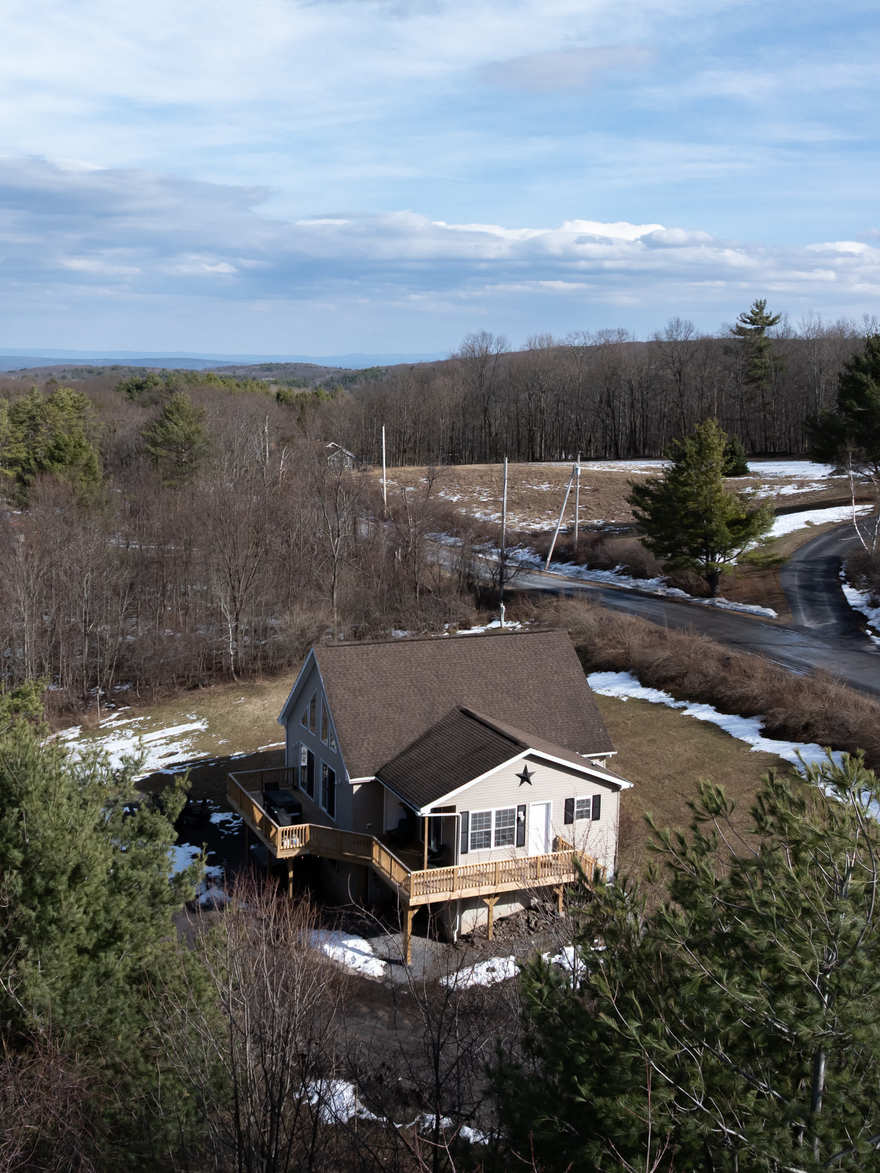 An aerial perspective reveals a modern house with a brown roof and light siding, surrounded by an expansive wooden deck. The property is set amidst sparse winter trees and patches of snow, with distant mountains under a partly cloudy sky.