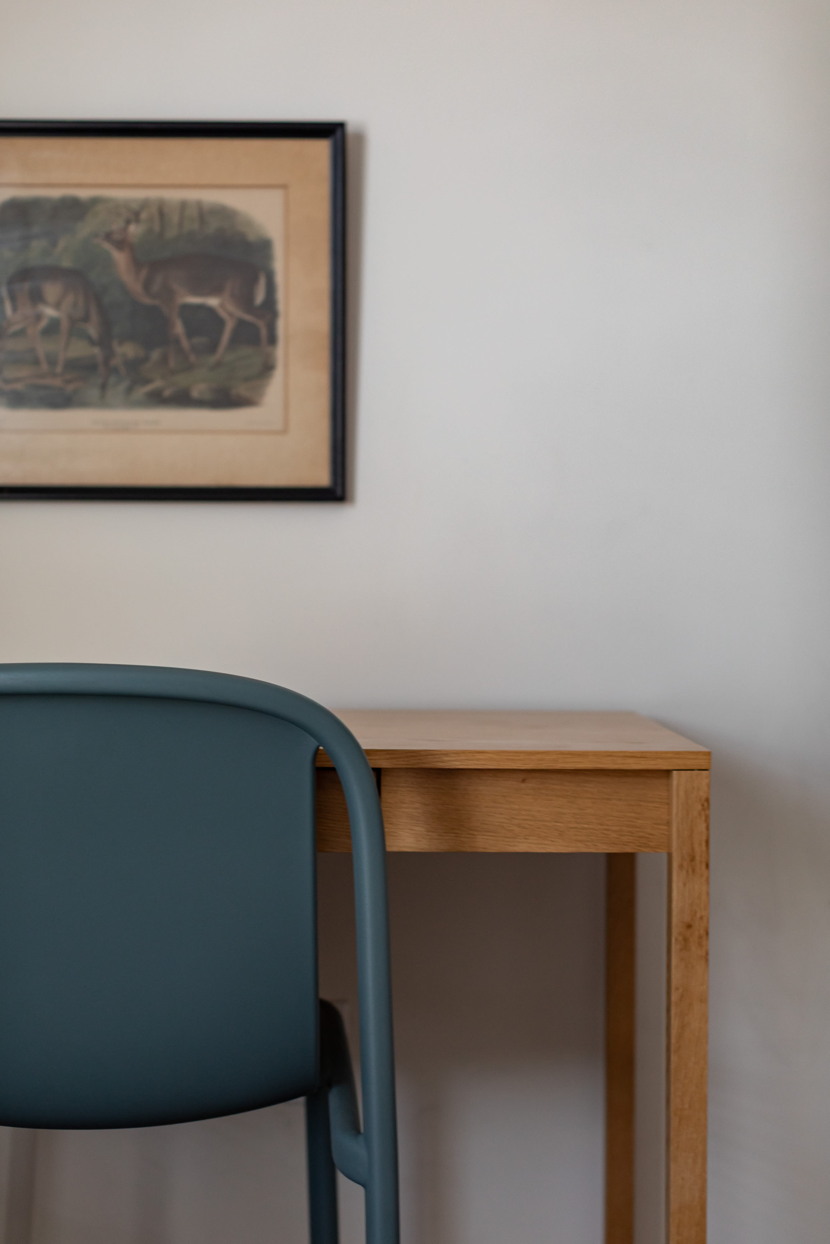 A minimalist workspace featuring a light wood desk and a dark teal chair, with a framed vintage-style print of deer on the white wall above, suggesting a quiet area for work or study.