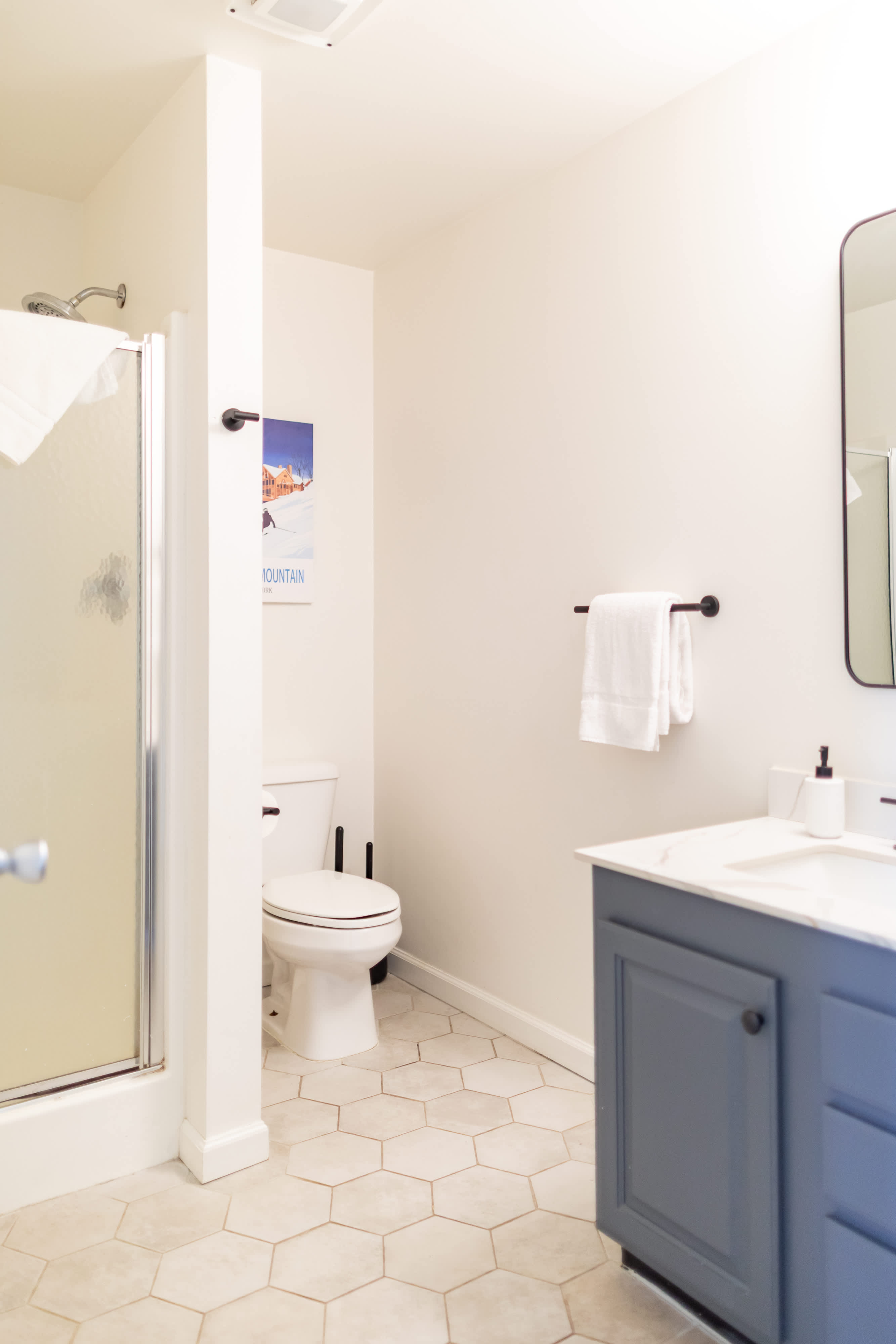 A modern bathroom featuring light hexagonal floor tiles, a glass-enclosed shower, a white toilet, and a vanity with a blue cabinet, white countertop, and a black-framed mirror. White towels are neatly folded.