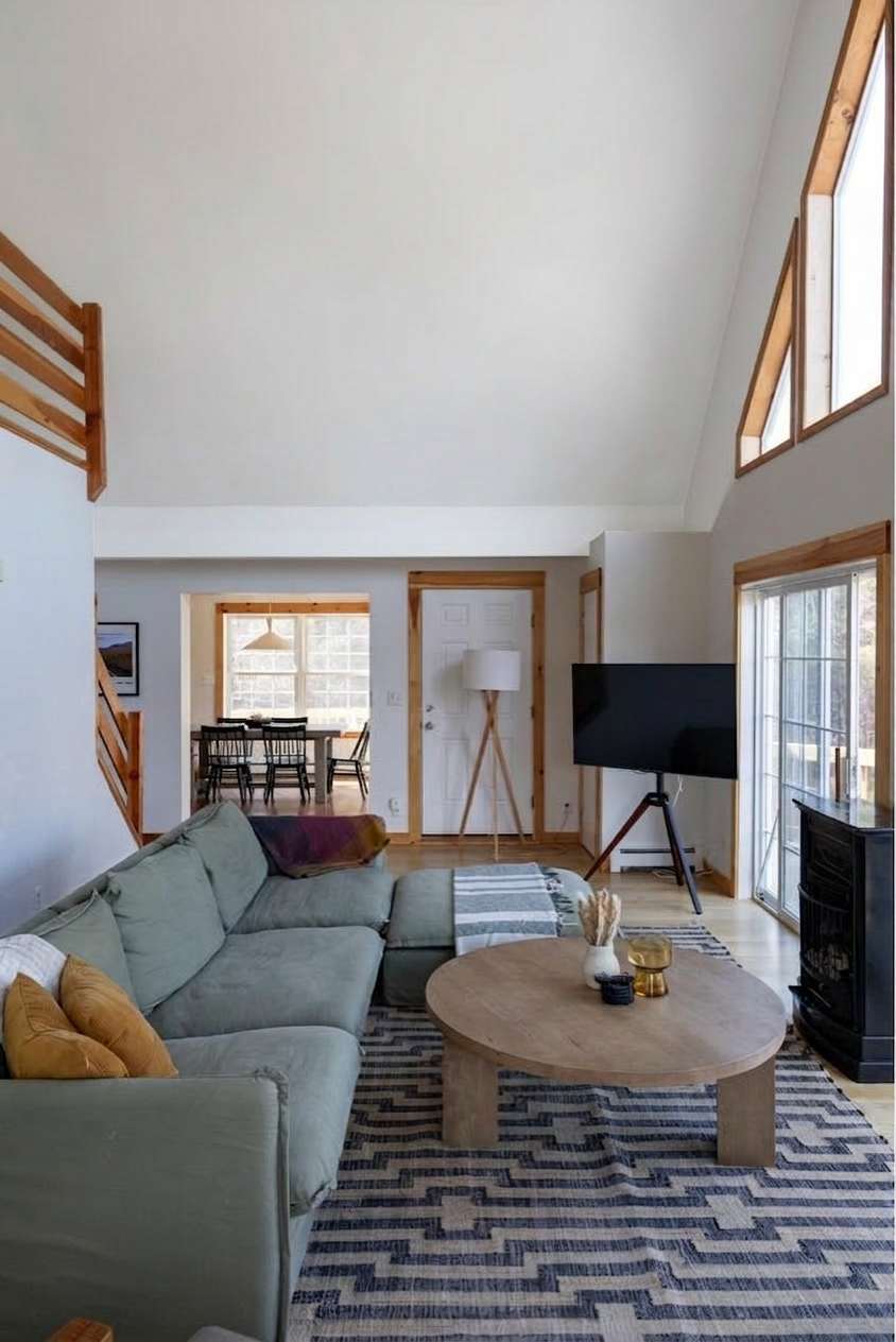 An expansive living room with vaulted ceilings and light wood floors features a large L-shaped green sectional sofa, a round wooden coffee table, and a wall-mounted TV on a tripod stand. A blue and white geometric rug anchors the seating area, with a view into the dining room beyond.