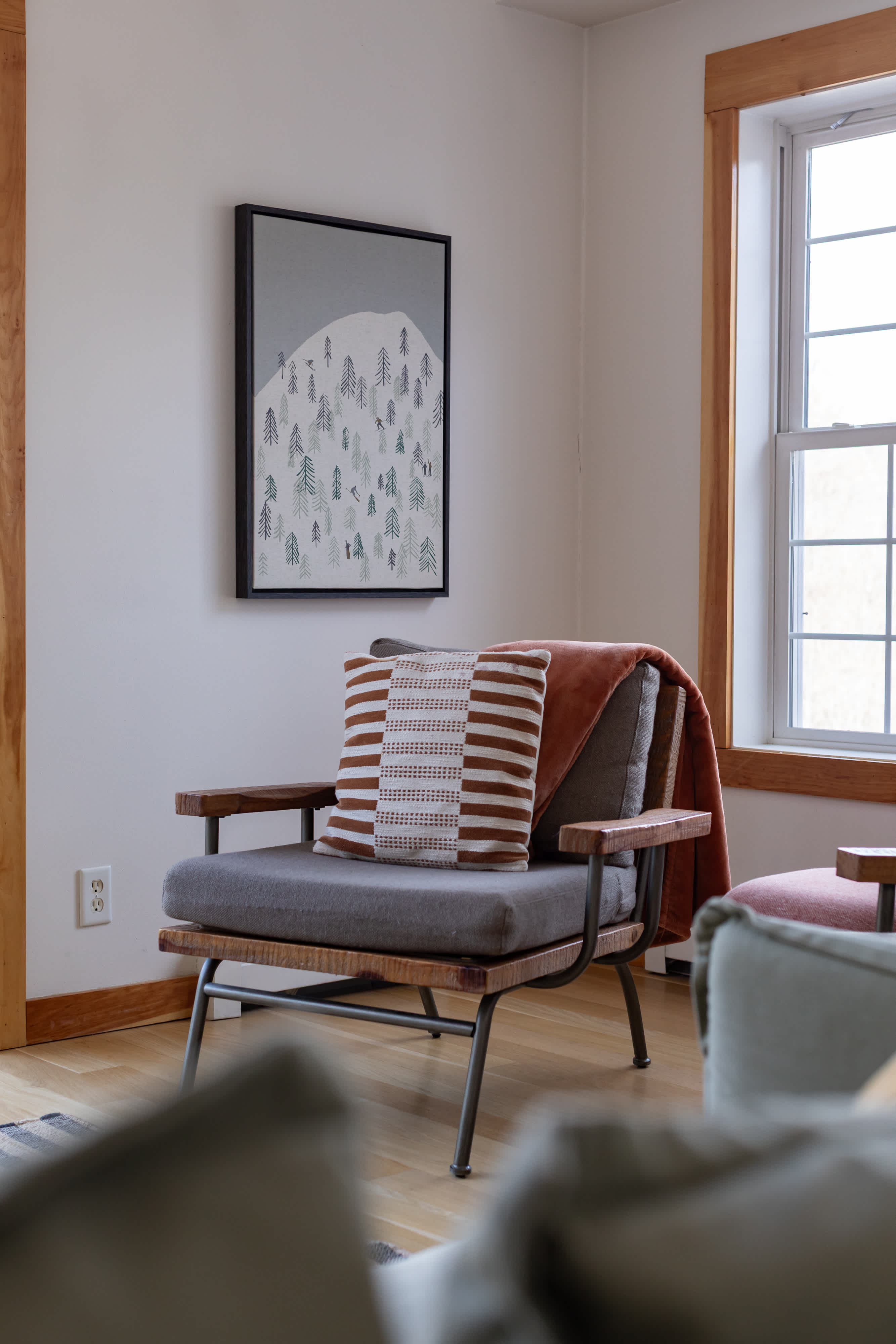 A close-up of a stylish gray armchair with wooden arms and metal legs, adorned with a striped pillow and a rust-colored throw blanket, positioned next to a large window with natural wood trim and a framed mountain art print on the wall.