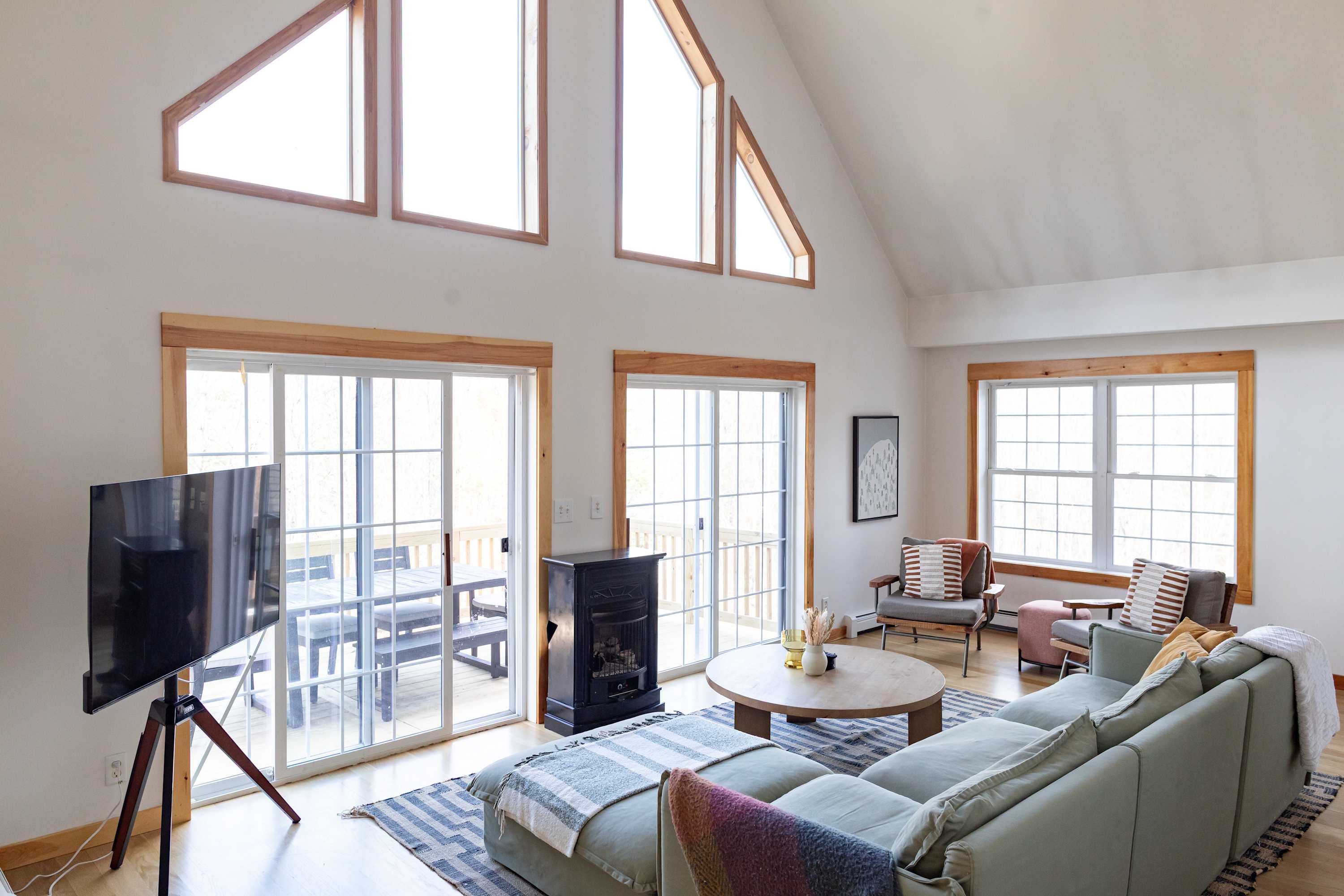 A bright and airy living room boasts a large light green sectional sofa, a round wooden coffee table, and a TV on a stand. Large windows and sliding glass doors offer abundant natural light and views, complemented by a small black fireplace and vaulted ceilings.