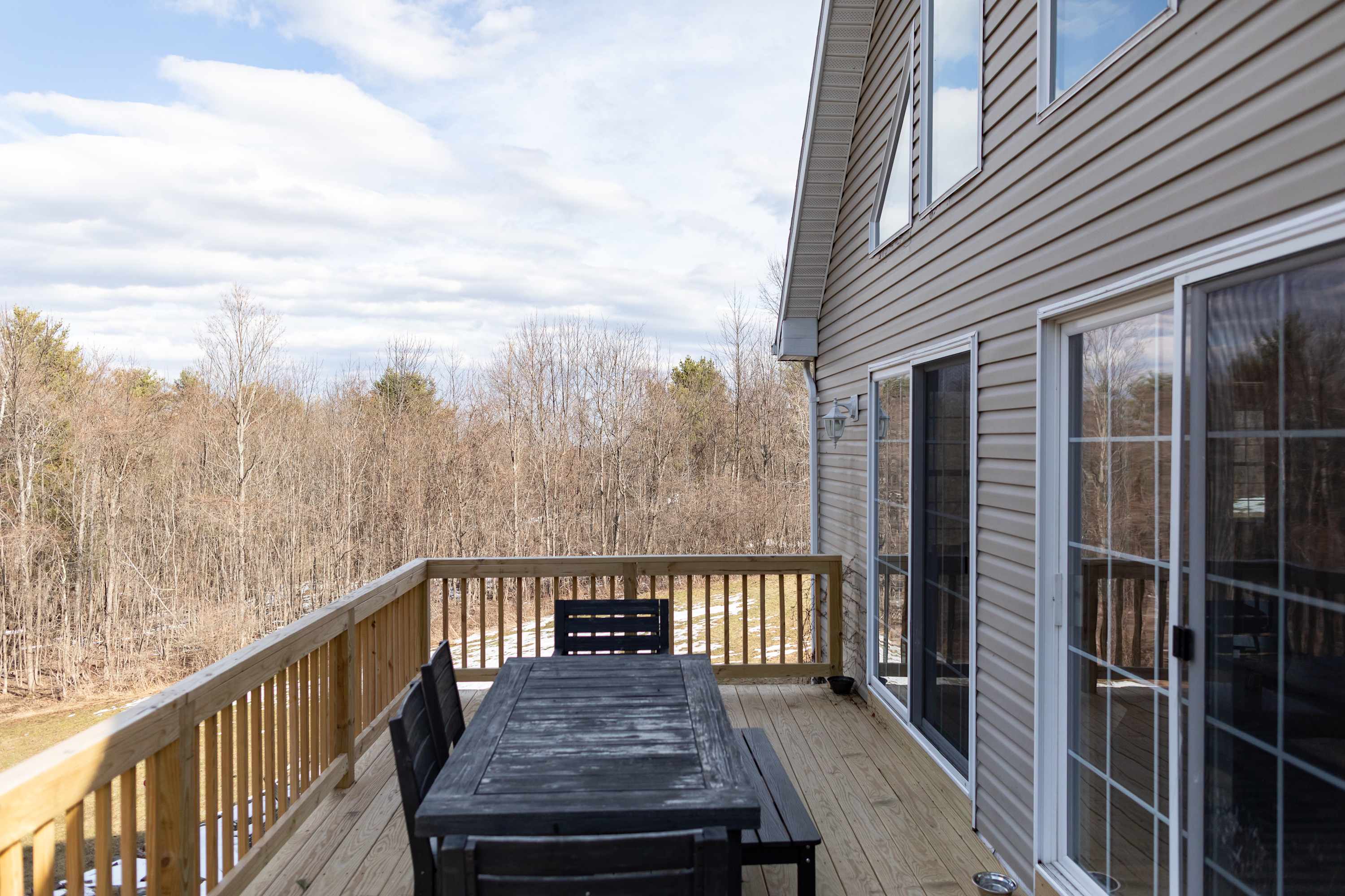 A spacious wooden deck features a large dark outdoor dining table with matching chairs, offering an inviting space for al fresco meals. It overlooks a winter landscape of bare trees and patches of snow, with the house's light siding and sliding glass doors visible.
