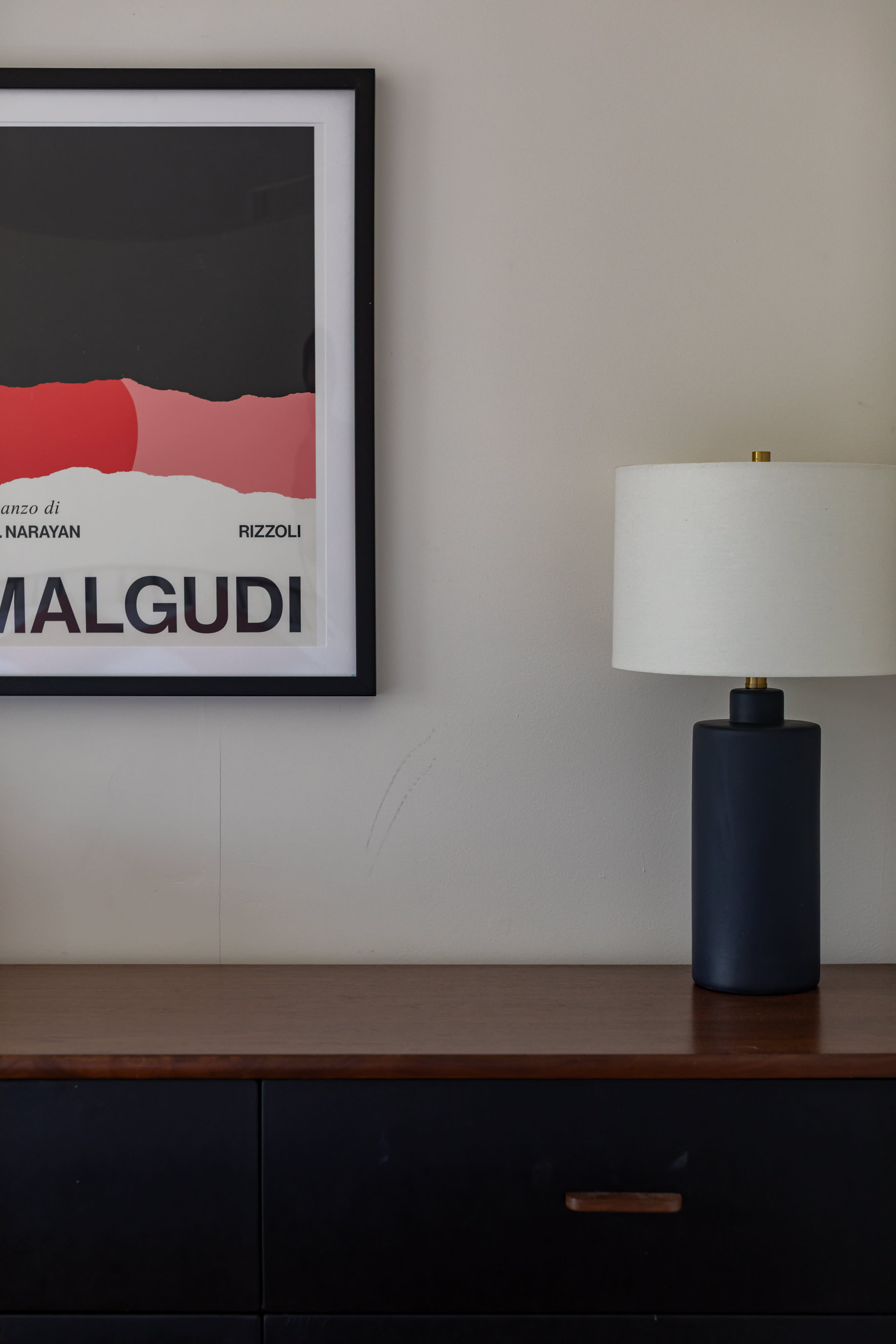 A close-up of a dark wood top dresser with black drawers and pulls, featuring a modern black table lamp with a white shade. Above, a framed abstract art print with red and black elements hangs on a light beige wall.