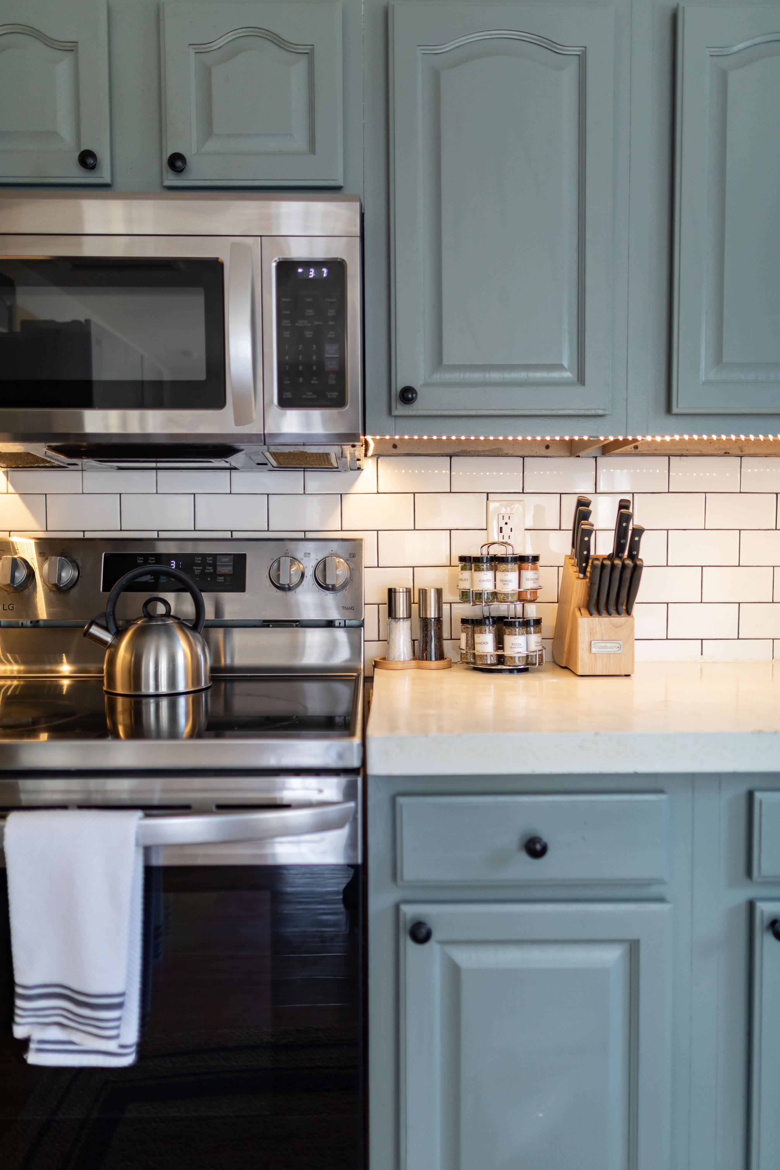 A close-up of a modern kitchen counter area, showcasing a stainless steel electric range with a kettle, a microwave above, white subway tile backsplash, light blue cabinets, and a collection of spices and knives.