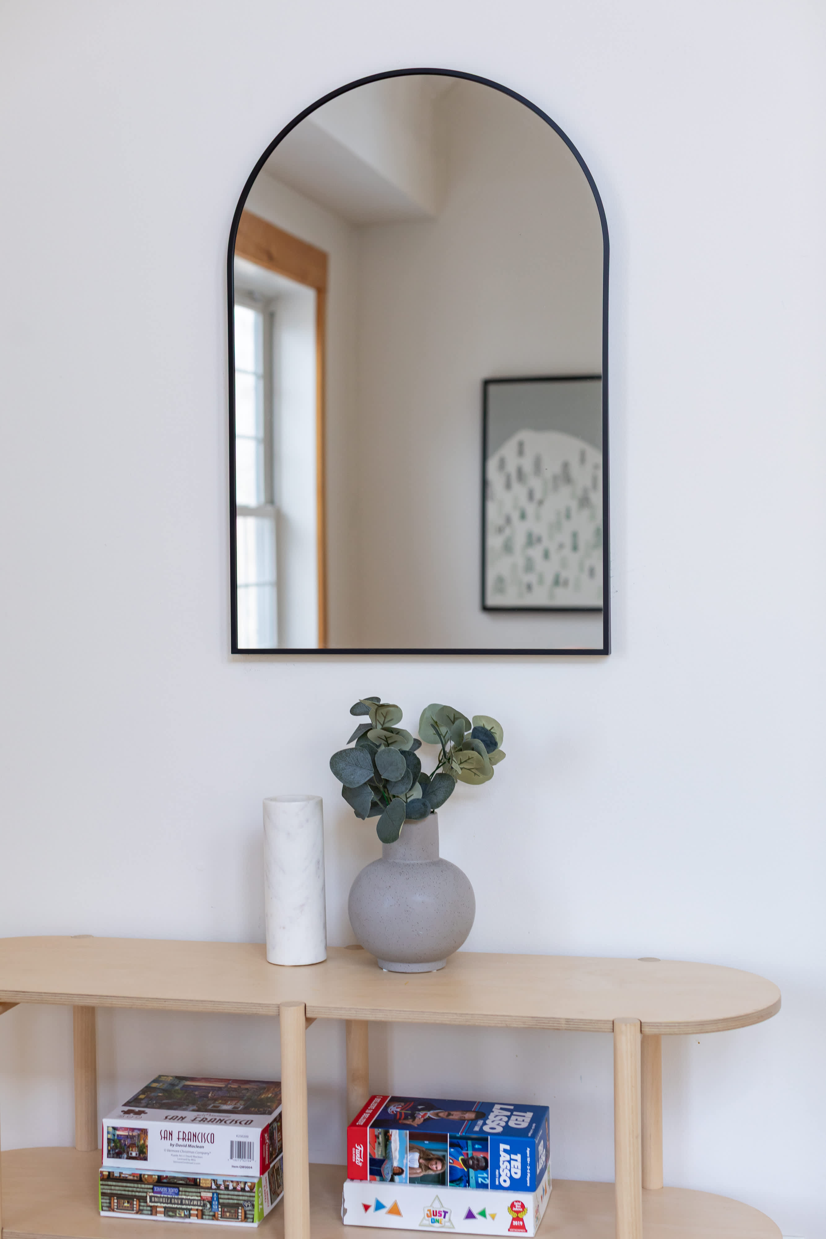 A light wood console table with open shelving holds board games, positioned beneath an arched black-framed mirror. A grey textured vase with artificial eucalyptus and a white marble cylinder are displayed on the table.