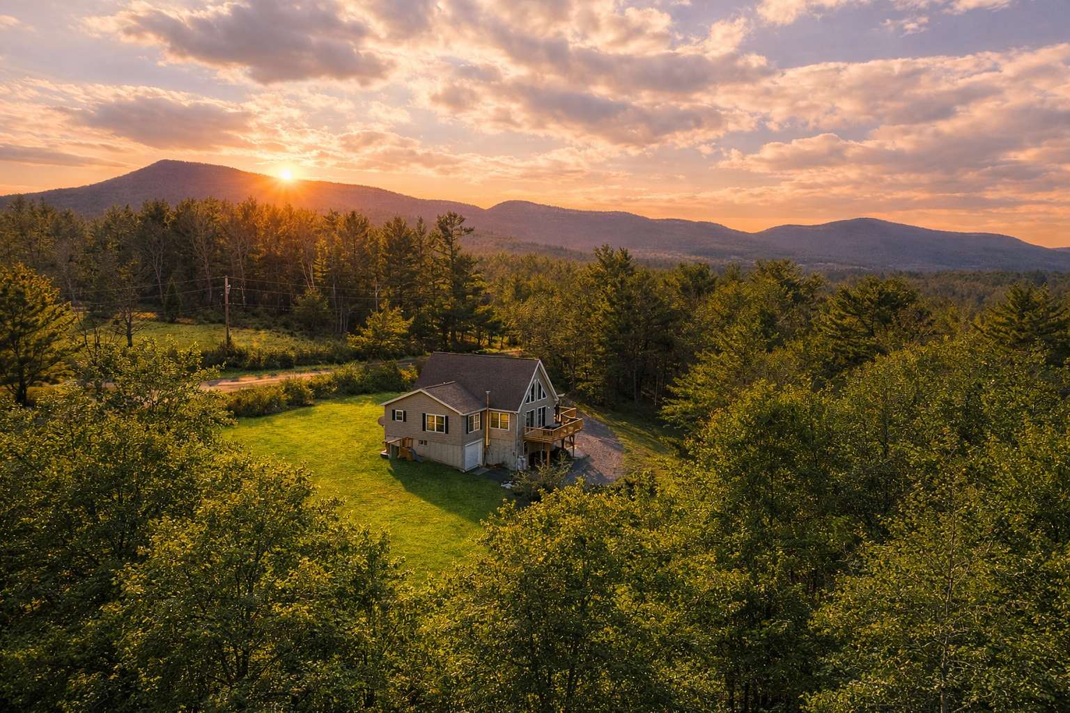 An aerial view of a modern house with a light gray exterior and a dark roof, nestled in a lush green clearing surrounded by dense forest, with a vibrant sunset casting warm light over distant mountains.