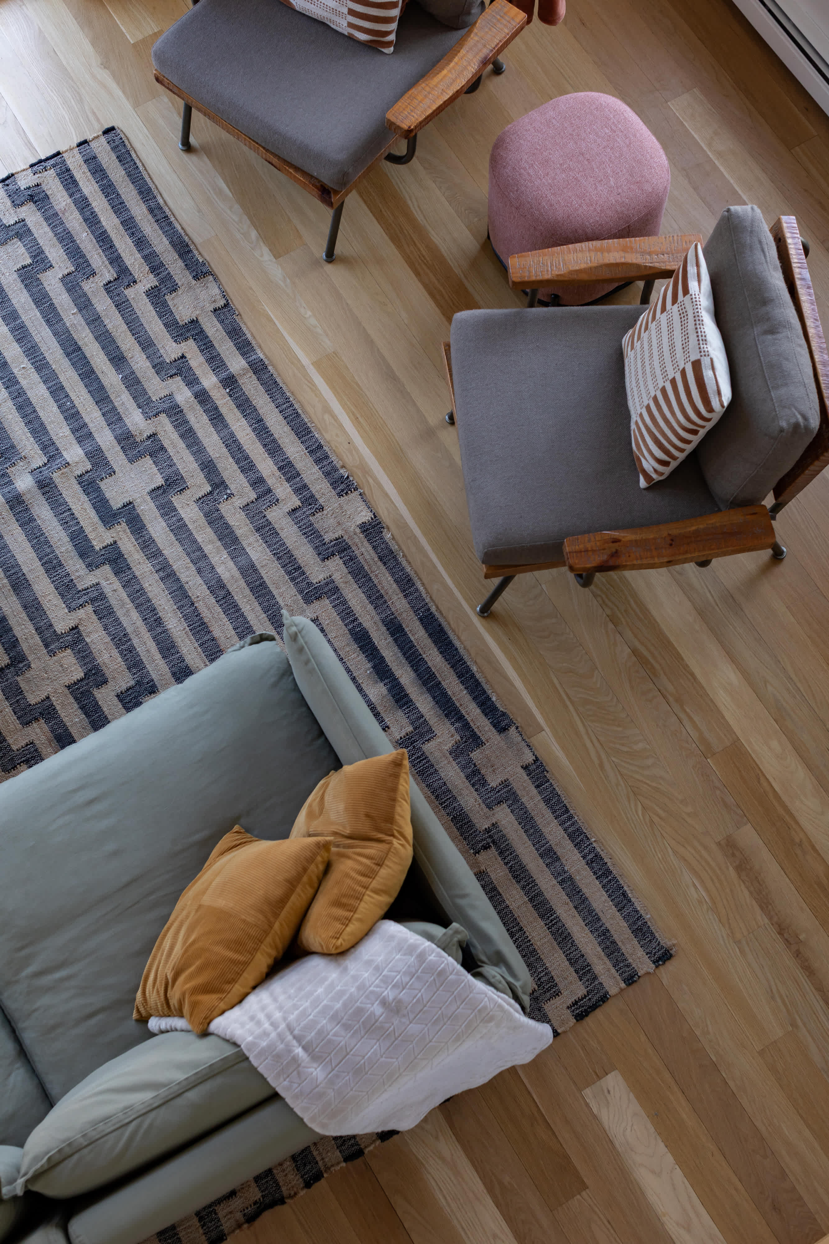 An overhead view captures a cozy living area with a light green sectional sofa, two grey armchairs, and a pink ottoman. The furniture is arranged on a striped area rug over light hardwood floors, adorned with various throw pillows.