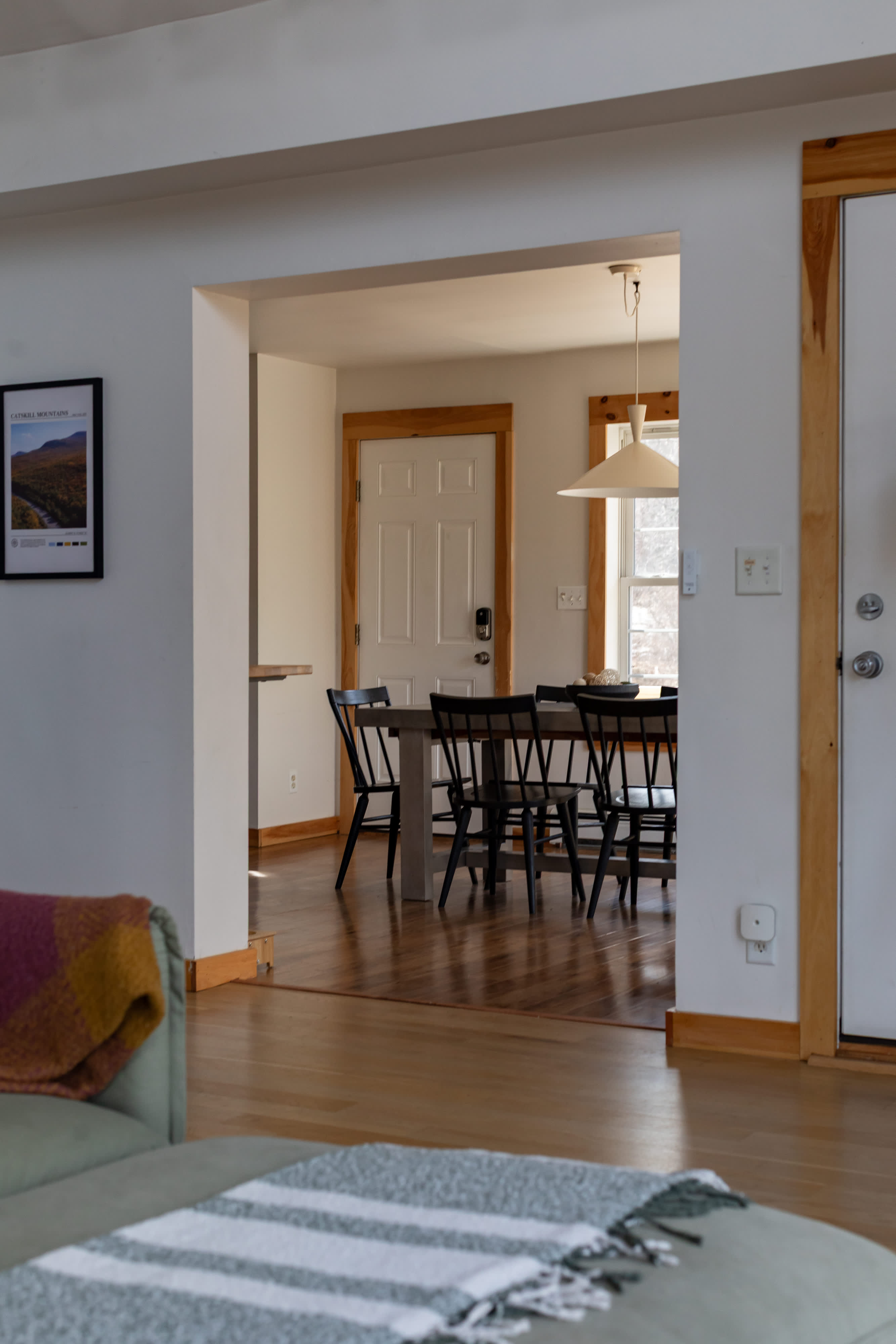 A view from the living area into a bright dining room, featuring a dark dining table surrounded by black chairs and illuminated by a white pendant light. A framed poster adorns the wall, and a white door is visible in the background.