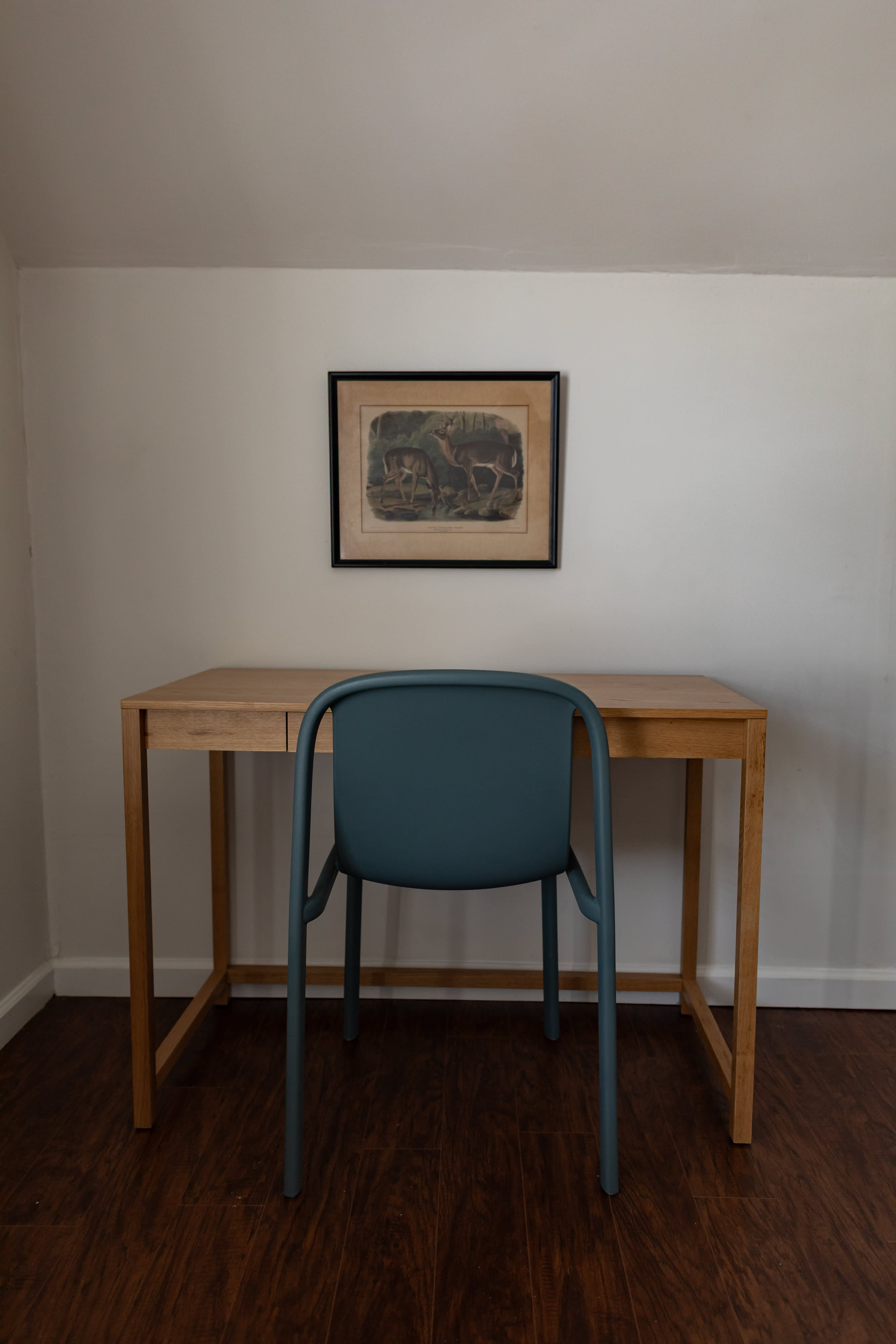 A minimalist workspace features a light wooden desk and a modern teal chair, set against a clean white wall. A framed nature print hangs above the desk, complementing the dark wood flooring.