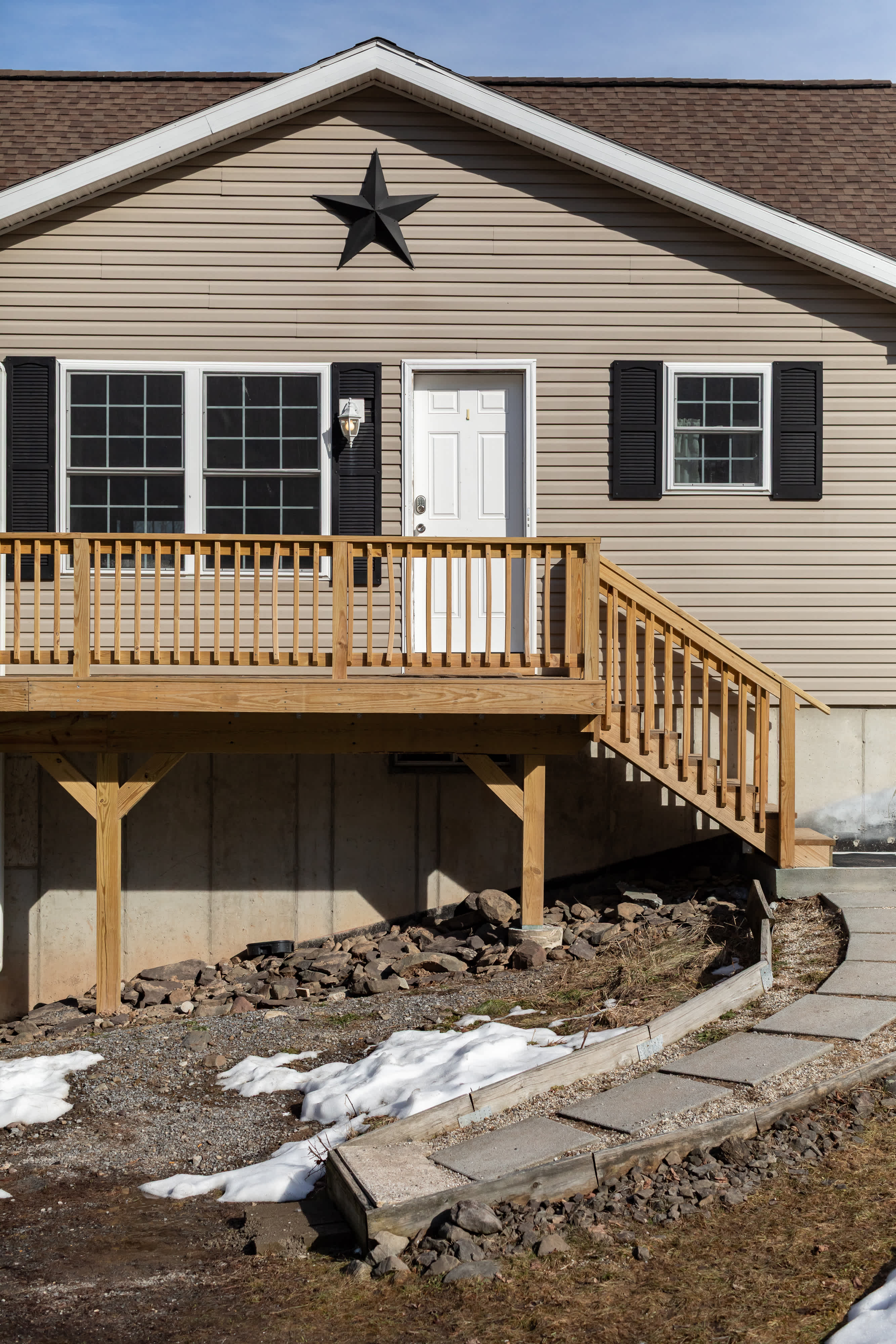 The front exterior of the house showcases light beige siding, black shutters, and a white front door adorned with a black star decoration above. A welcoming wooden deck with stairs leads up to the entrance, with patches of snow on the ground.