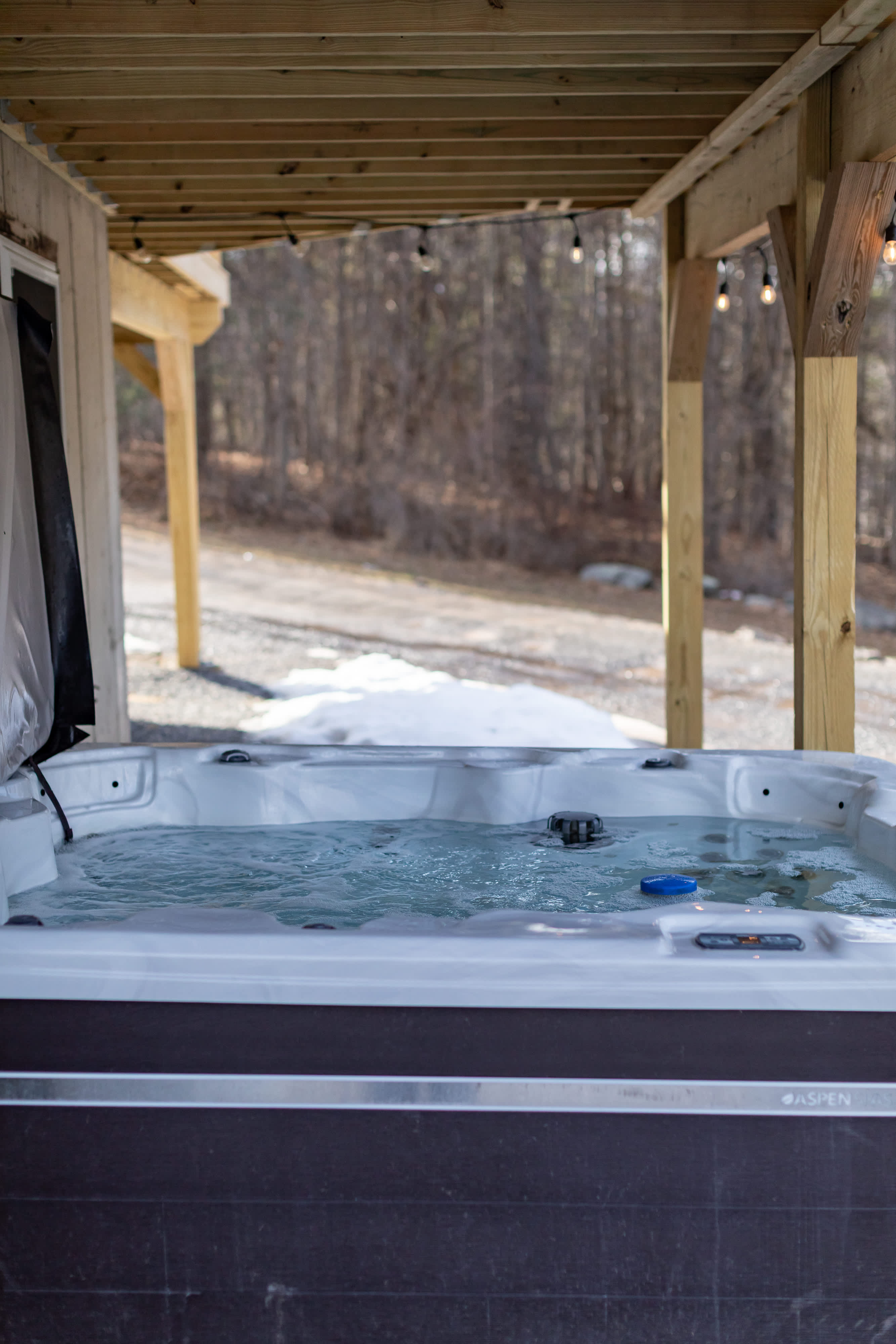 A bubbling hot tub is nestled beneath a wooden deck, illuminated by string lights, offering a relaxing spot with a view of a snowy, wooded landscape. The dark grey hot tub features a blue cover accessory.