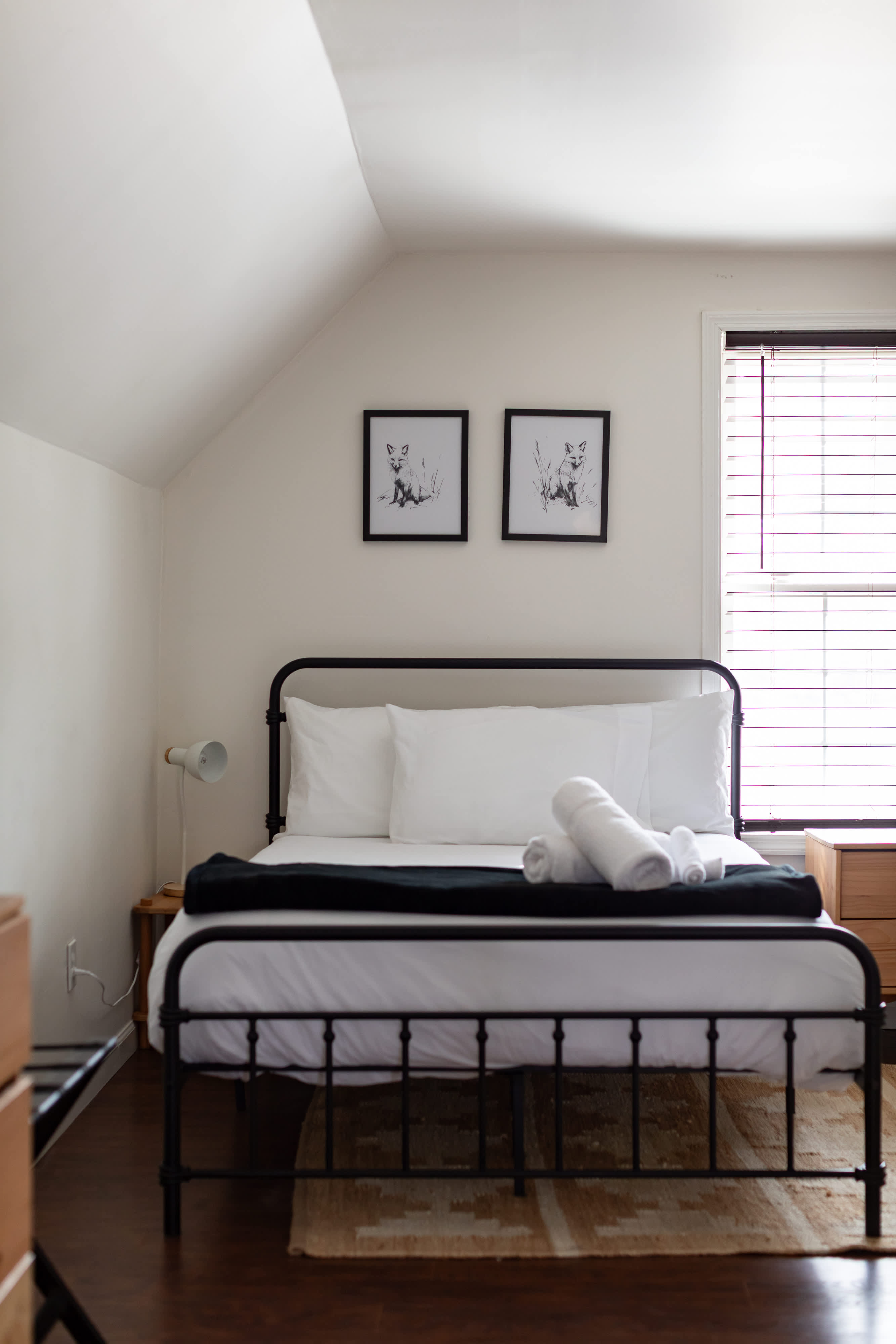 A cozy bedroom with a black metal bed, white bedding, and a black throw, set against a sloped ceiling. Two framed black and white fox prints hang above the bed, next to a window with blinds.