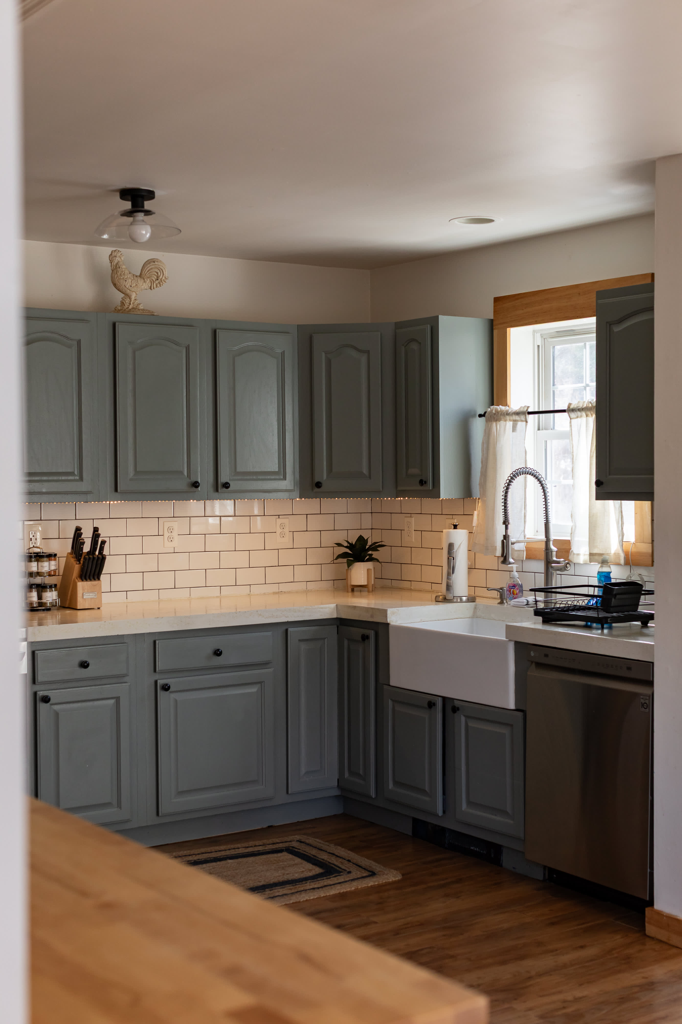 A U-shaped kitchen features sage green shaker-style cabinets with black hardware, white subway tile backsplash, and light countertops. A white farmhouse sink with a gooseneck faucet is positioned under a window with sheer curtains, and stainless steel appliances are visible.