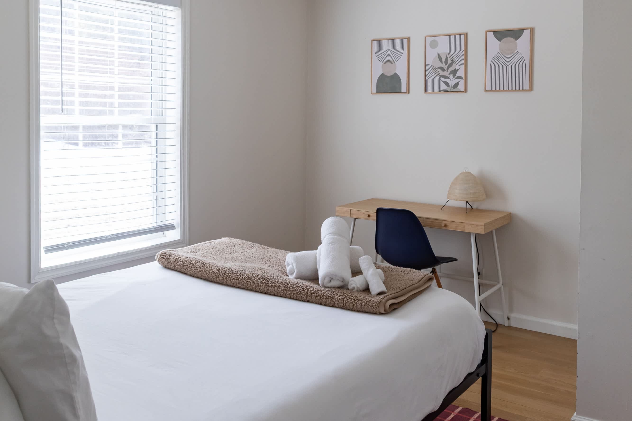 A detailed view of a wooden desk with a woven table lamp and a dark blue chair in a bright bedroom. Three framed abstract prints hang above the desk, complementing the light-colored walls and window with blinds.