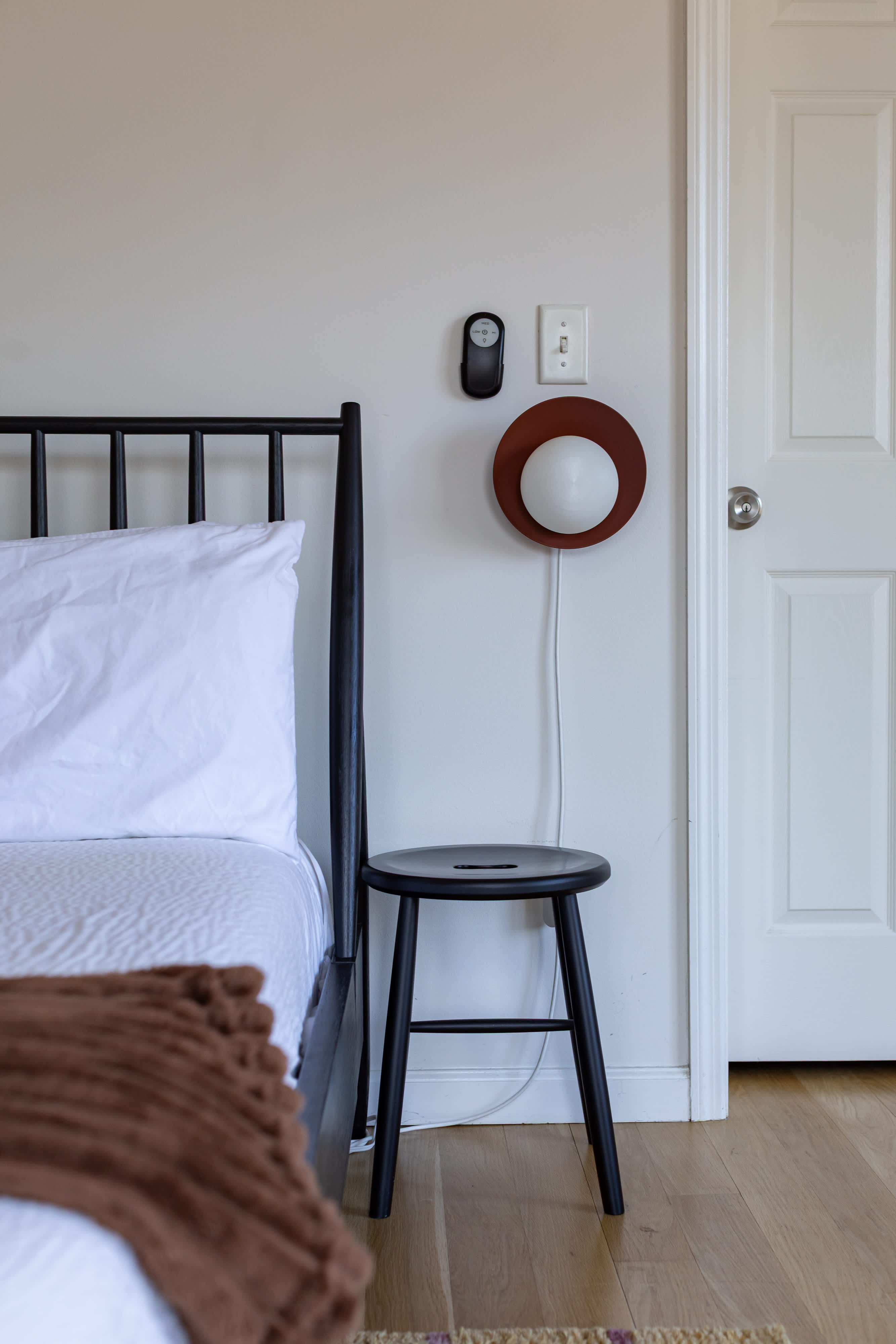 A close-up of a black metal bed frame with white bedding and a brown throw, next to a black wooden stool. A modern circular wall sconce and a smart thermostat are mounted on the light-colored wall.