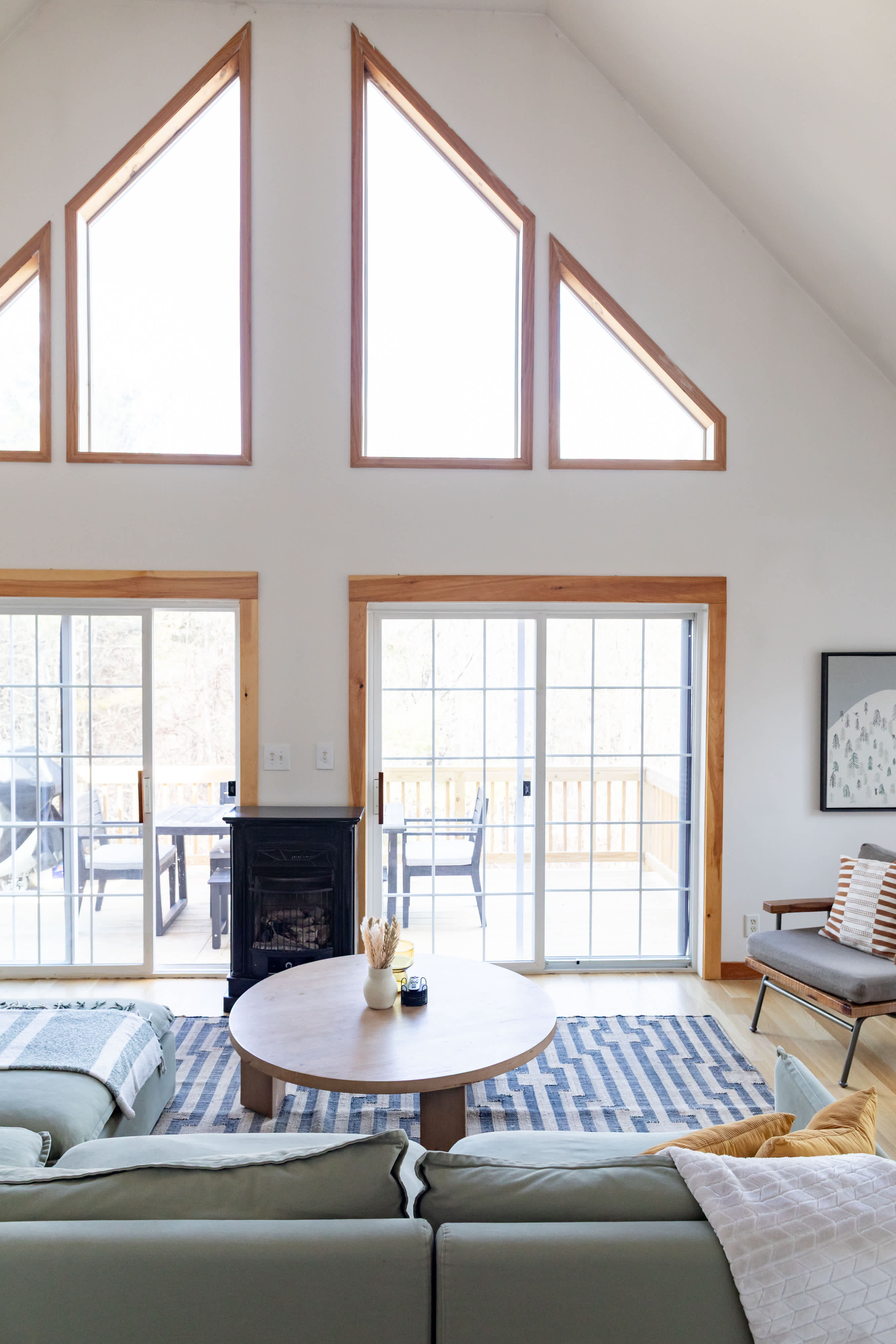 A bright living room with vaulted ceilings and light wood floors features a large L-shaped green sectional sofa and a round wooden coffee table on a blue and white geometric rug. Multiple large windows and sliding glass doors with natural wood trim offer abundant natural light and views of the outdoors.