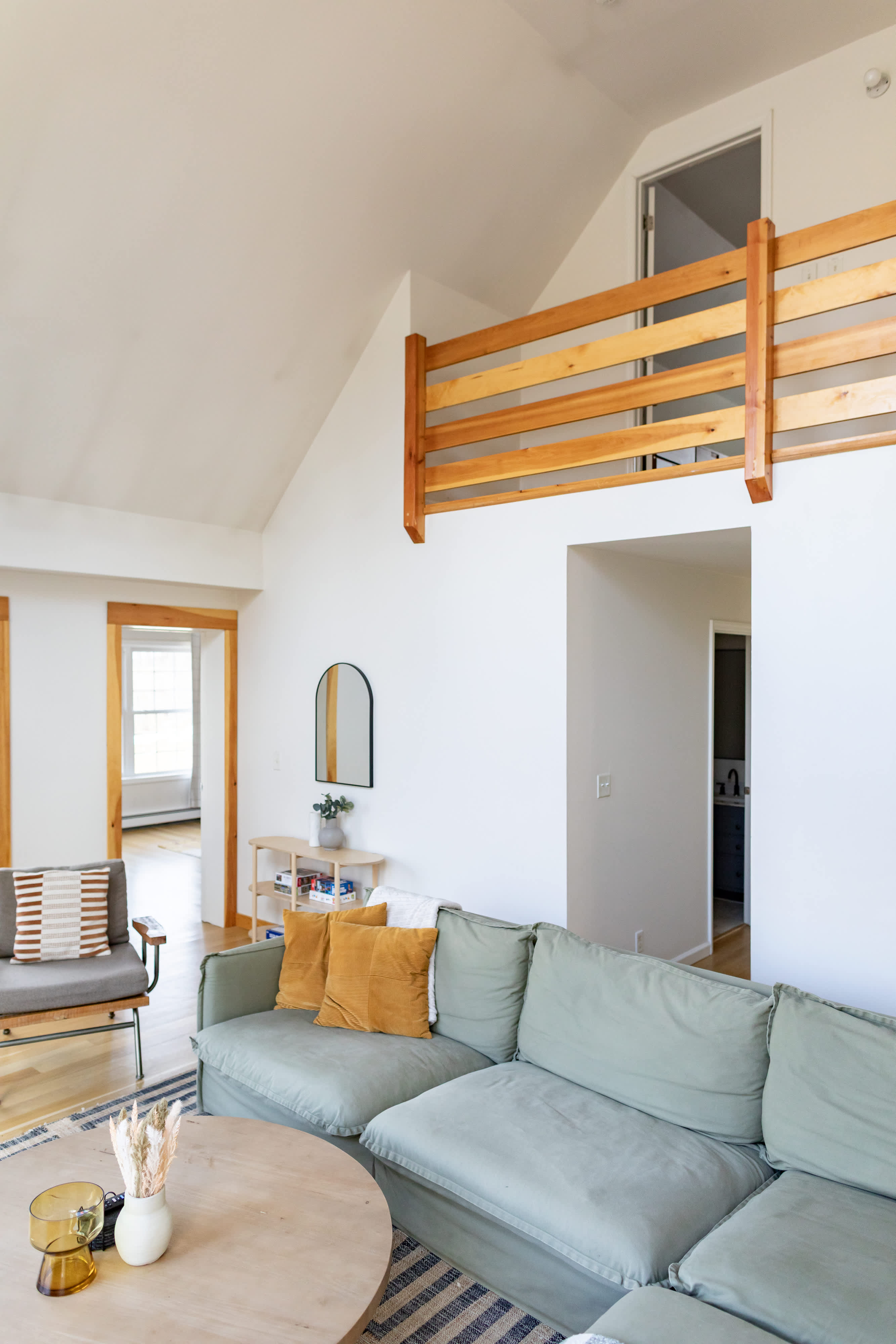 A bright living room features a large light green sectional sofa with mustard throw pillows and a round wooden coffee table. A wooden railing for a loft overlooks the space, with a hallway leading to other rooms.