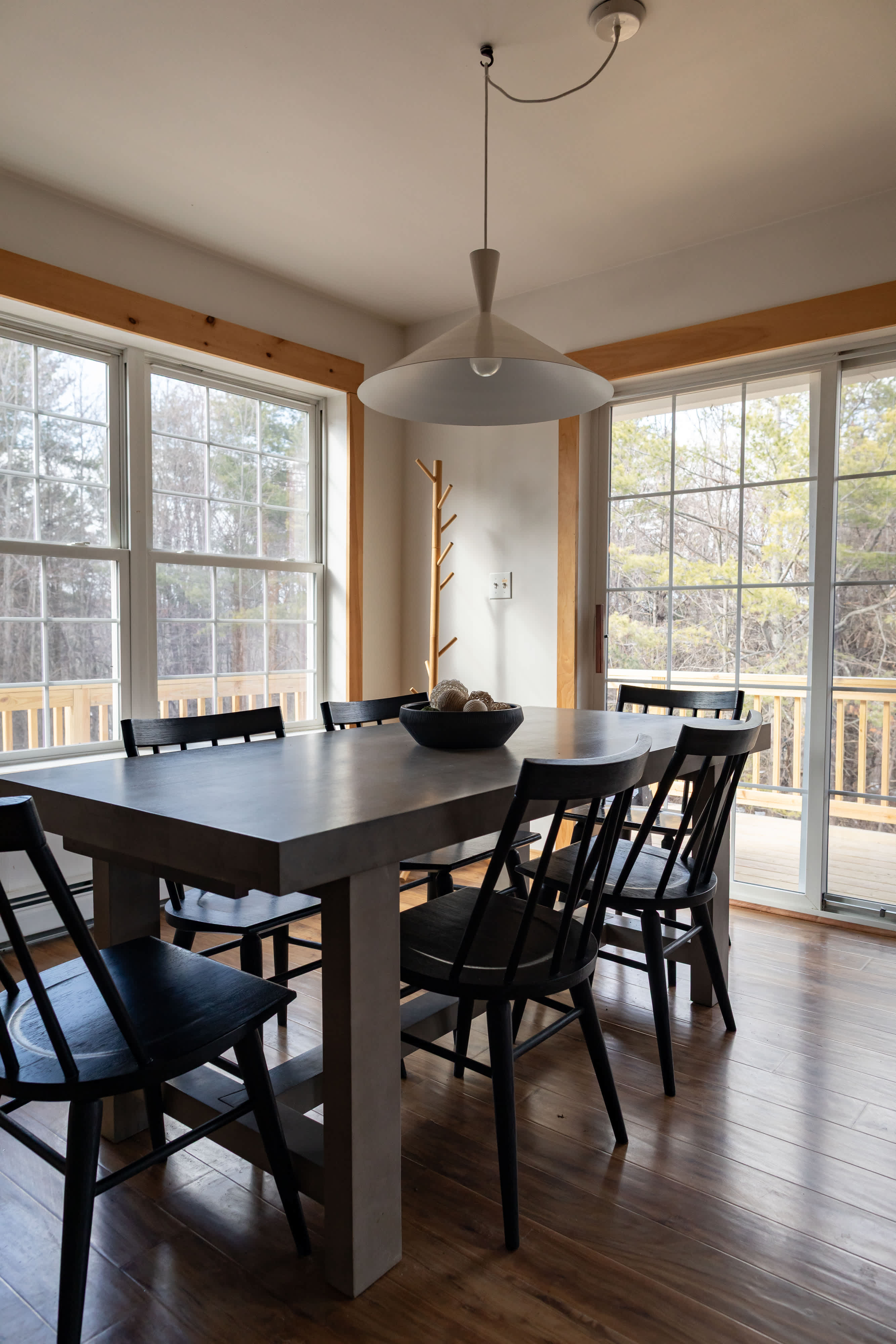 A spacious dining area features a large grey rectangular table surrounded by black spindle-back chairs, centered under a modern white pendant light. Sliding glass doors and large windows with natural wood trim offer views of an expansive outdoor deck and surrounding trees.