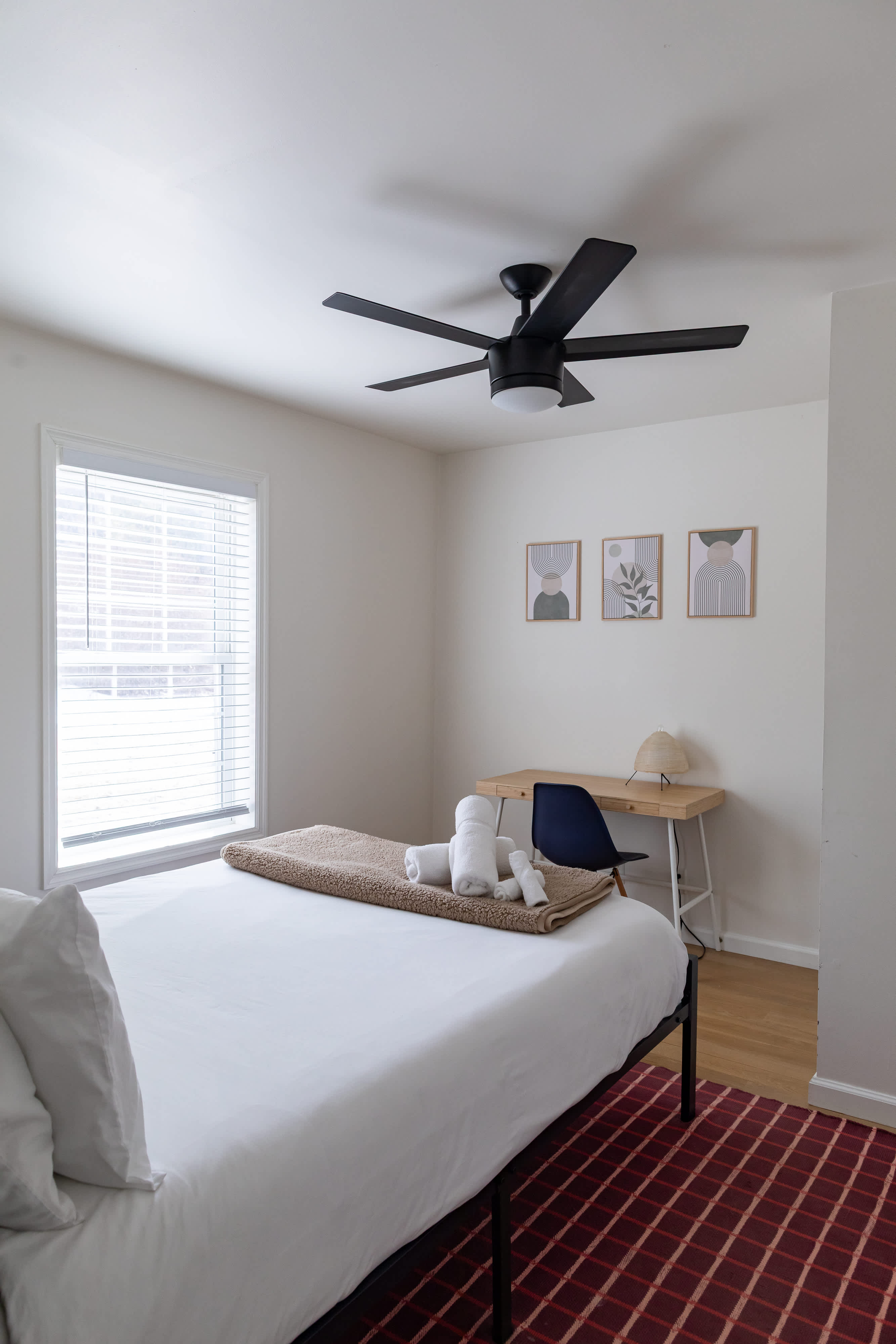 A bright bedroom featuring a black metal bed with white bedding and a brown textured throw, with a wooden desk and a dark blue chair in the corner. Three framed abstract prints adorn the wall above the desk, next to a window with blinds.