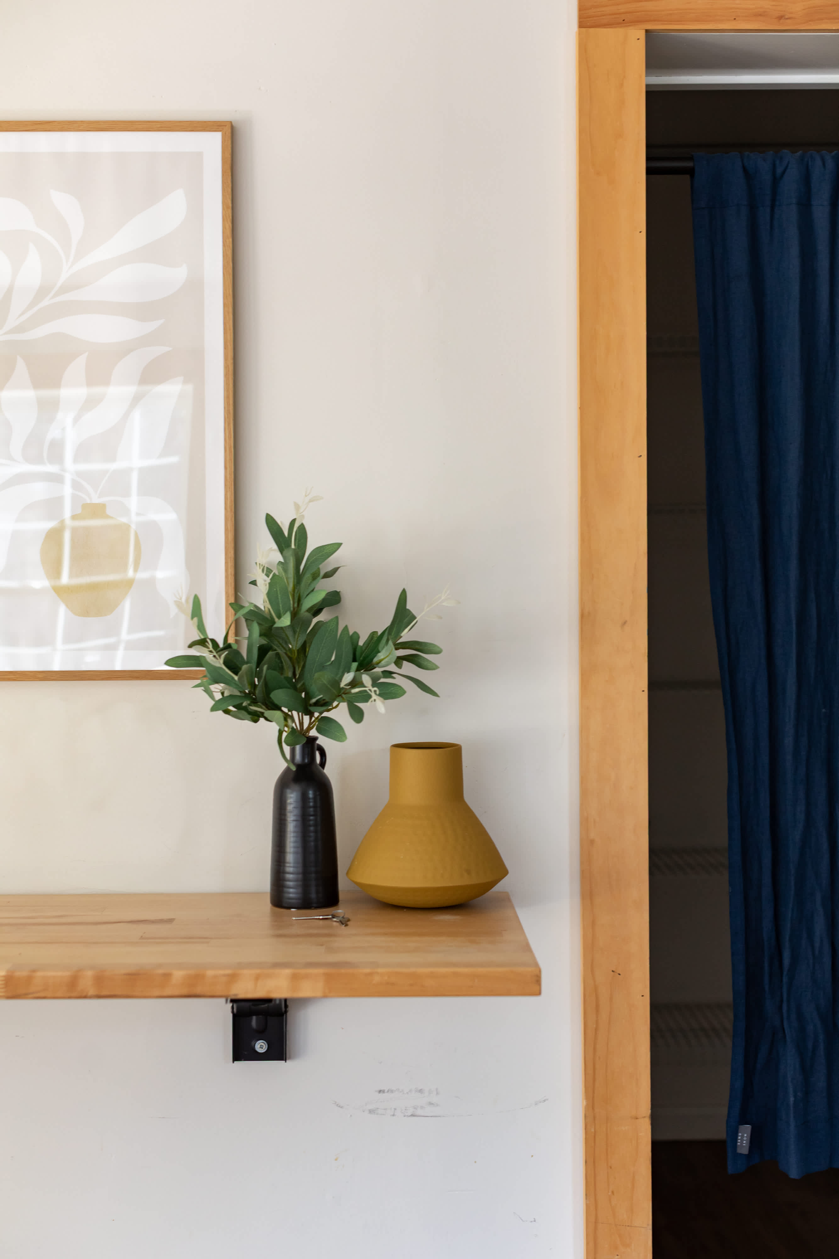 A light wood floating shelf against a light beige wall holds a black vase with artificial greenery and a mustard yellow decorative vase. Above, a framed abstract art print with a plant motif hangs, and to the right, a closet opening with a blue curtain is visible.