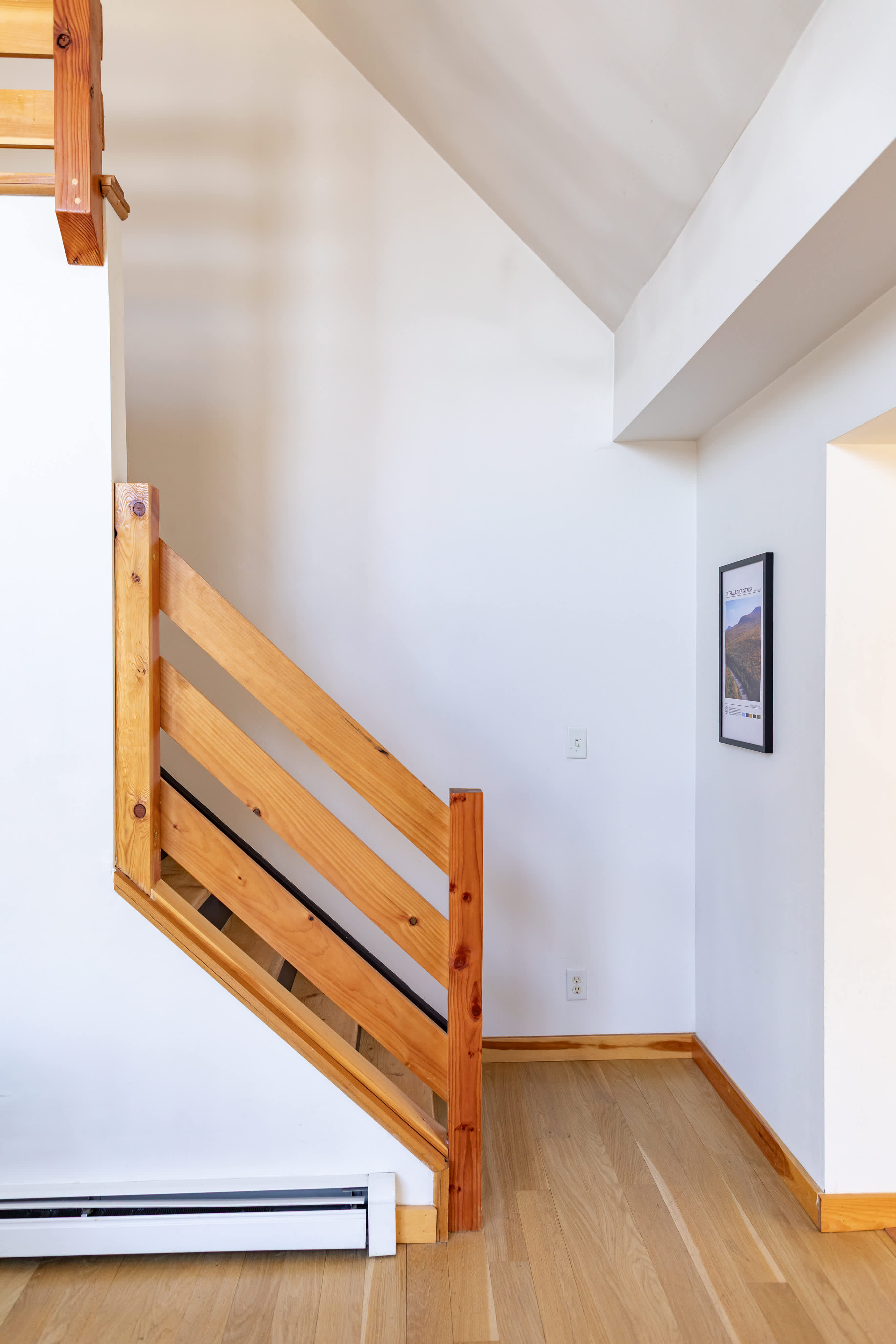 A bright hallway or entryway featuring a light wood staircase with horizontal railings, leading upwards. Light hardwood floors and white walls create an airy feel, with a baseboard heater visible.