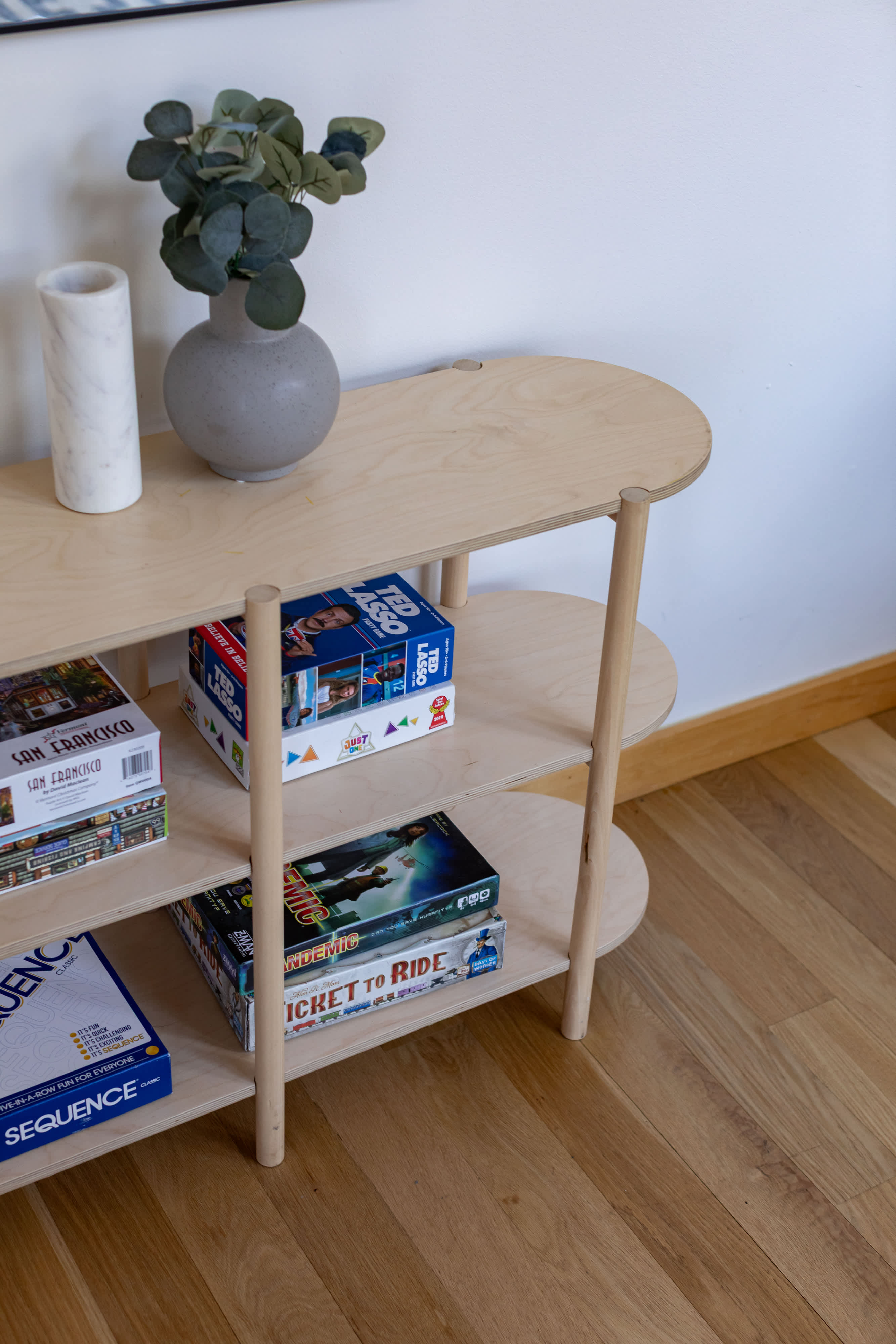 A light wood shelving unit displaying a collection of board games like "Ticket to Ride" and "Sequence," alongside decorative items such as a gray vase with eucalyptus and a marble holder, set against a white wall with light wood flooring.