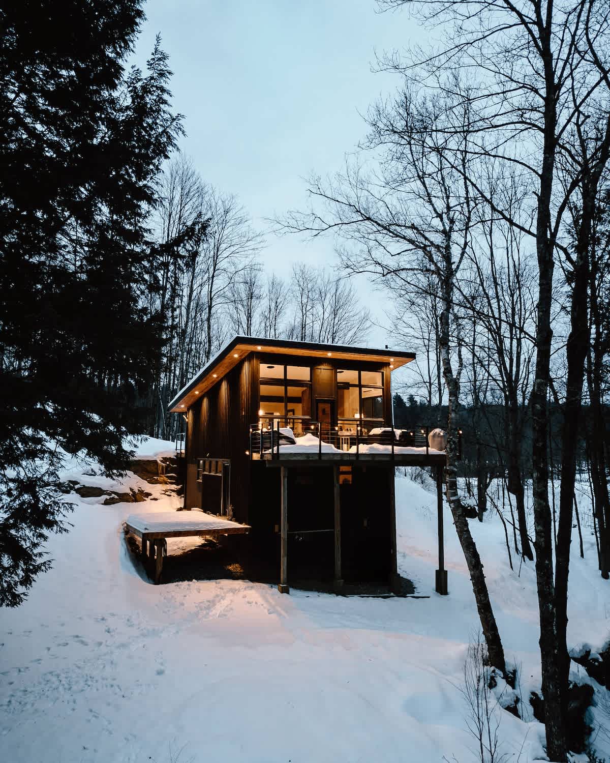 A modern black cabin with large windows and a raised deck is set in a snowy winter landscape, surrounded by bare trees and evergreens. Warm interior lights glow, creating a cozy contrast with the cold, serene environment.