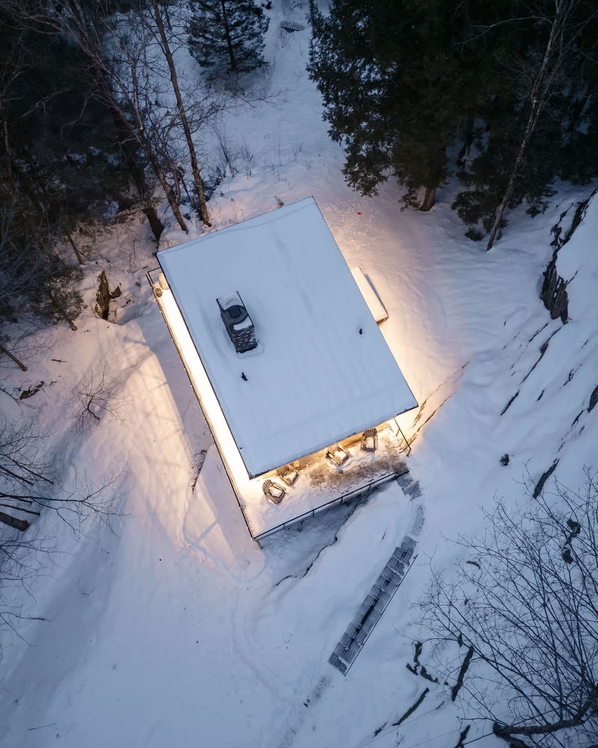Overhead aerial view of a modern house nestled in a snow-covered forest, featuring a flat roof, a prominent chimney, and illuminated eaves casting a warm glow on the surrounding snow and deck area.