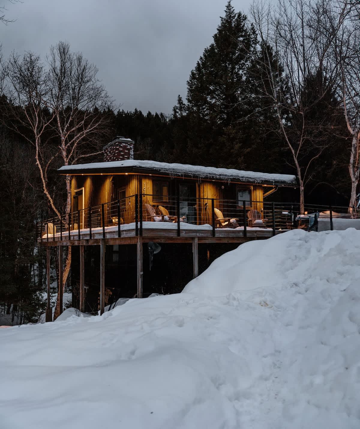 Elevated modern cabin with warm wooden siding, a stone chimney, and a spacious deck featuring outdoor seating, illuminated by string lights, all set against a snowy, forested backdrop at dusk.