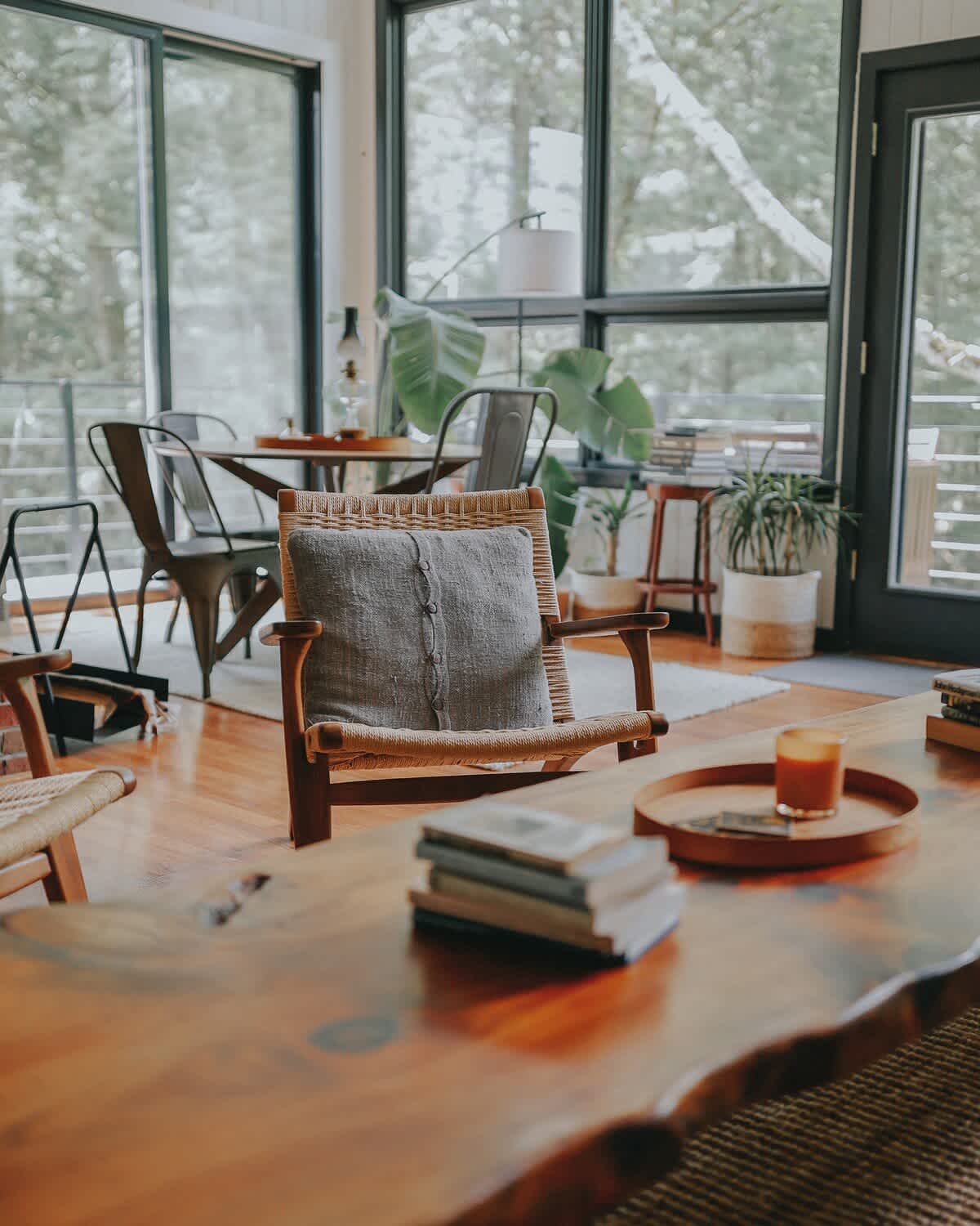 View from the living area showcasing a rustic live-edge coffee table and a woven armchair with a grey pillow, looking towards the dining area with its round table and metal chairs, all framed by expansive forest-facing windows.