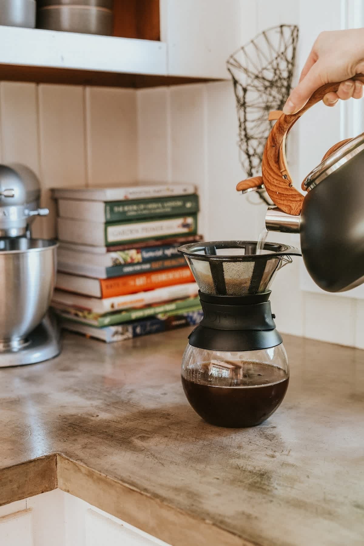 A close-up shot of a pour-over coffee brewing setup on a light-colored countertop, with a kettle pouring hot water into a glass carafe. Cookbooks and a stand mixer are visible in the background.