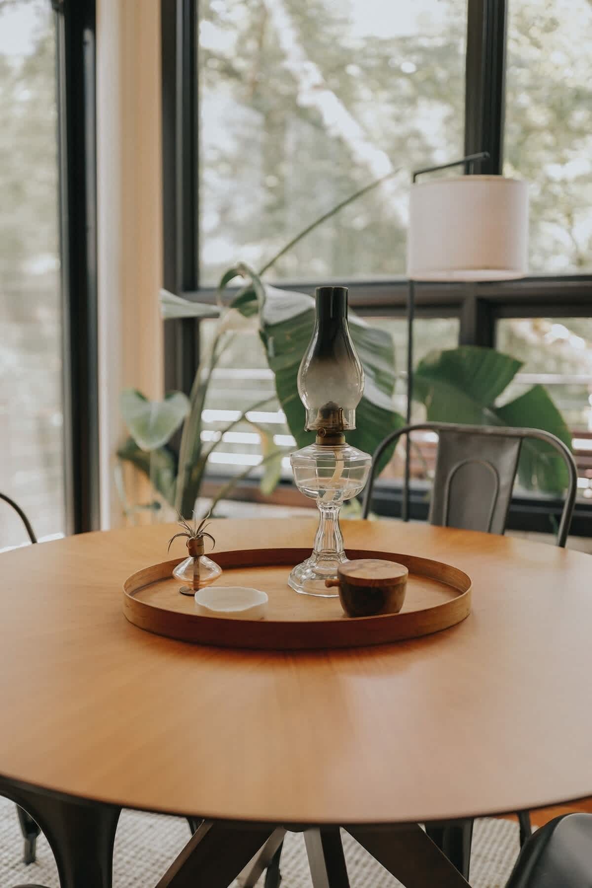 A close-up of the round wooden dining table, featuring a rustic wooden tray centerpiece with a vintage oil lamp, a small air plant, and a lit candle, against a backdrop of large windows.