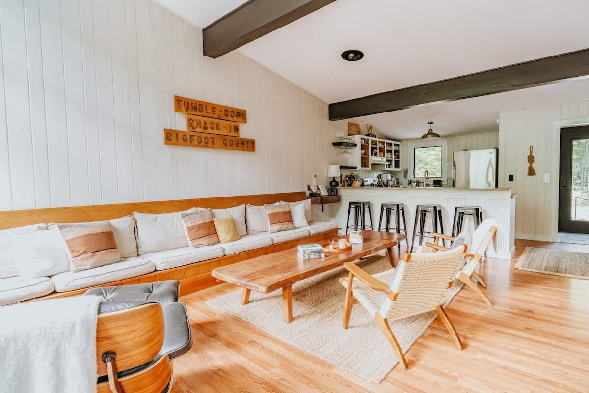 Spacious living room with a long, built-in wooden bench sofa adorned with white and earth-toned pillows, facing a large live-edge wooden coffee table. Beyond, a modern kitchen with a breakfast bar and stools is visible, all set on warm hardwood floors.
