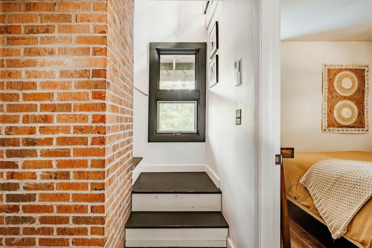 A narrow hallway or landing featuring a prominent exposed brick wall on the left, dark-stained wooden stairs, and a small window with dark trim. A doorway on the right reveals a glimpse of a bedroom.