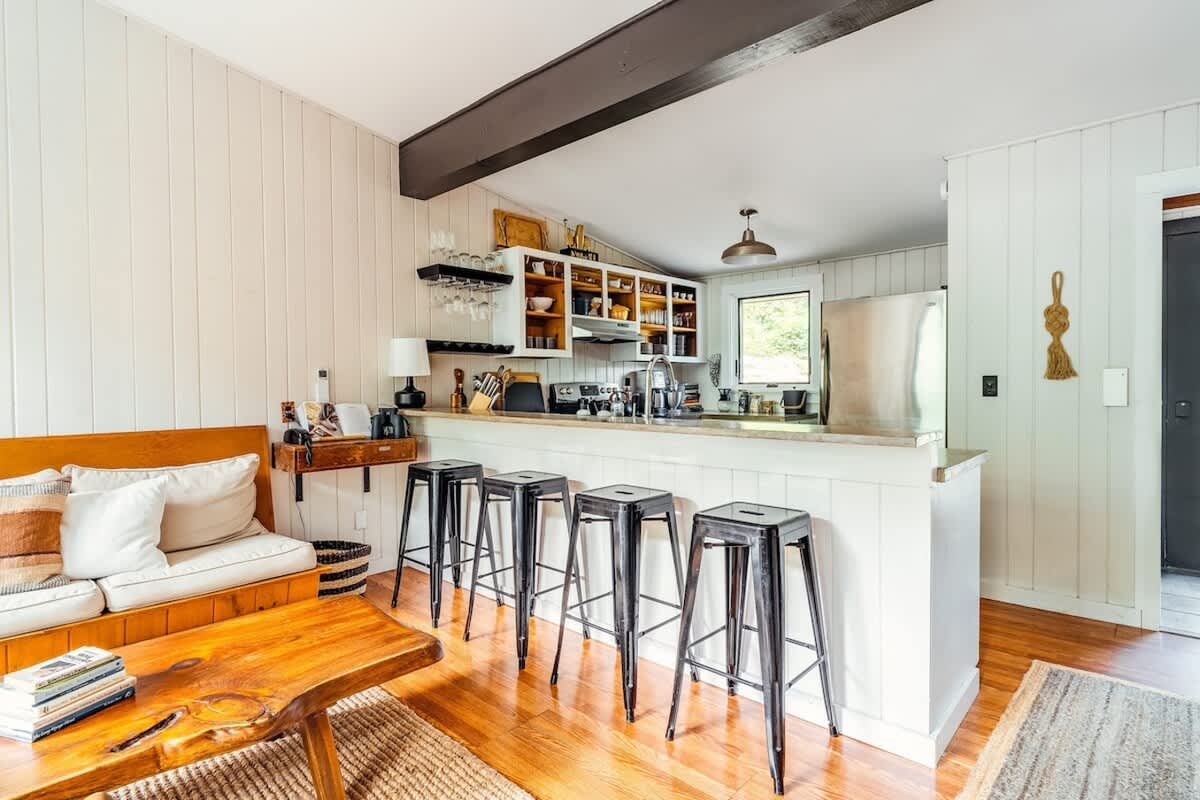Open-concept kitchen with a white breakfast bar featuring black industrial stools, concrete countertops, and white cabinetry with open shelving, seamlessly connecting to the living area.