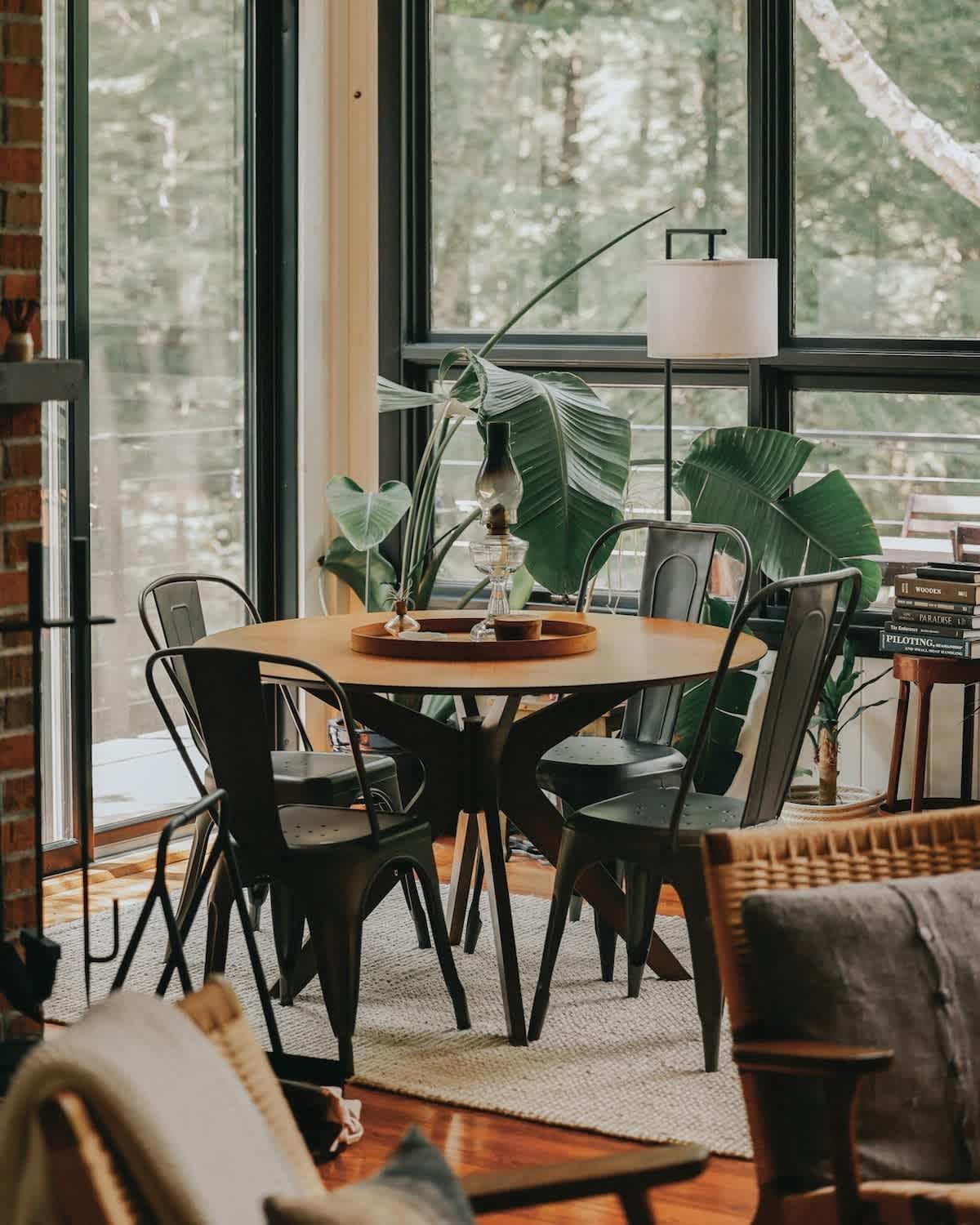 Cozy dining area featuring a round wooden table with four black metal chairs, set against large windows overlooking lush greenery, with a tall plant and a table lamp adding to the ambiance.