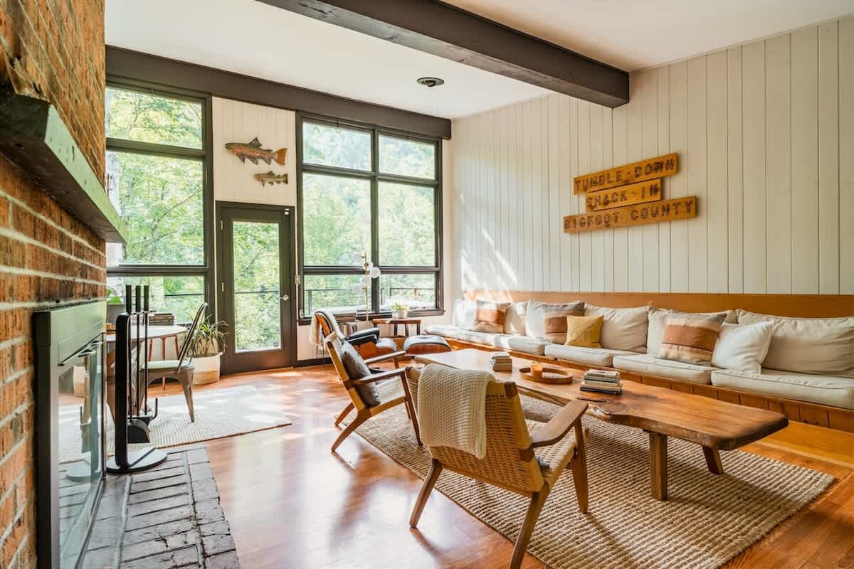 Expansive living room featuring a long built-in wooden sofa with cushions, large windows spanning the wall offering abundant natural light and forest views, and a rustic wooden coffee table on a jute rug.