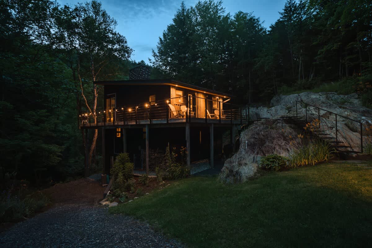 The modern black cabin is illuminated at dusk, with warm lights glowing from inside and string lights adorning the raised wooden deck. It is nestled on a rocky, grassy slope surrounded by dark forest, with a gravel driveway leading up to it.