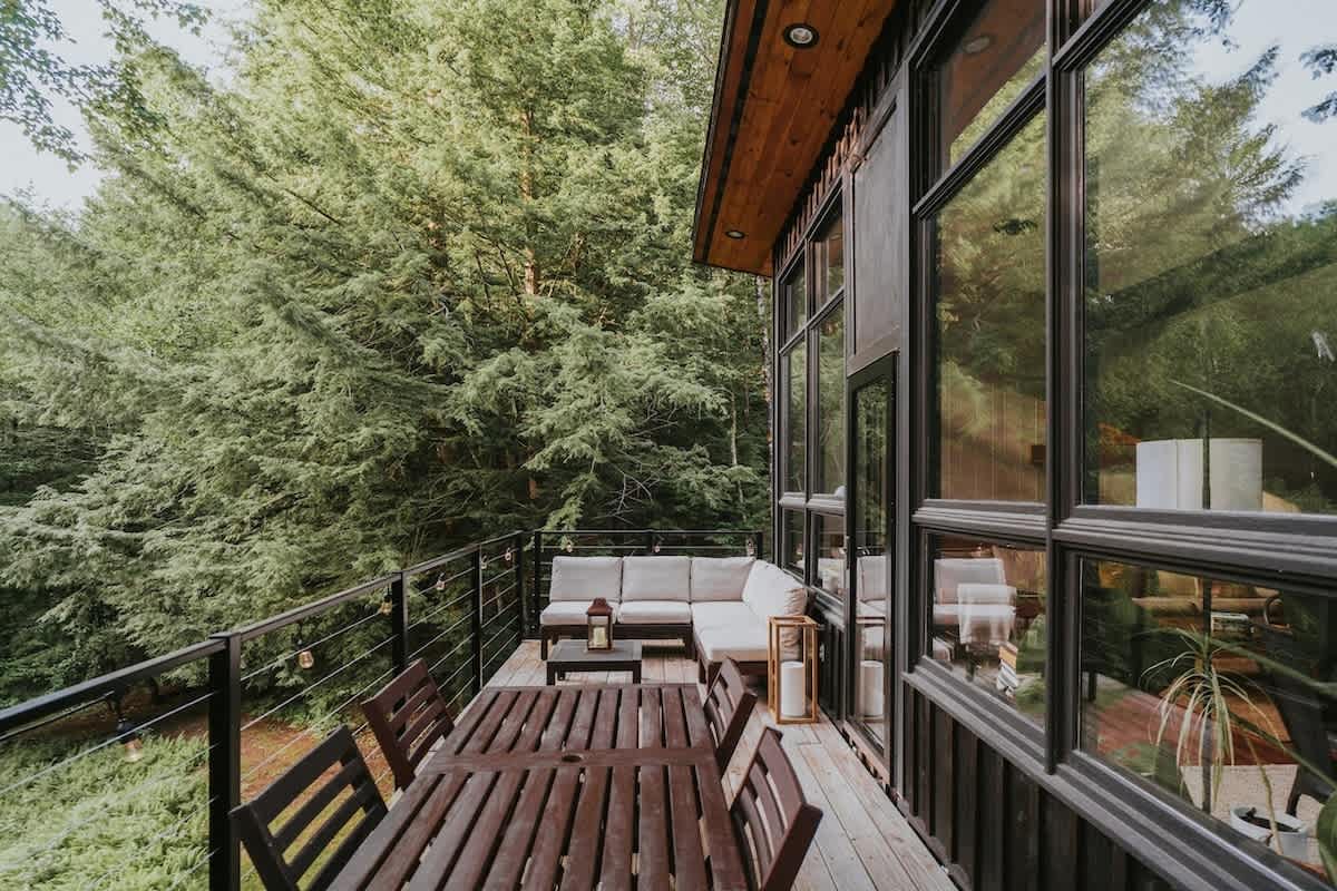 A wide wooden deck featuring an outdoor dining table and chairs, alongside a comfortable sectional sofa, all offering expansive views of the surrounding dense forest.