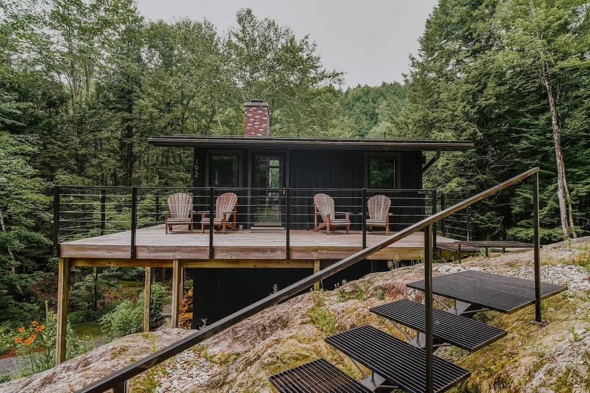 A modern black cabin with a flat roof and brick chimney is nestled among lush green trees, featuring a raised wooden deck with black metal railings and four Adirondack chairs. A metal staircase with open treads leads up to the deck from a rocky, grassy slope.
